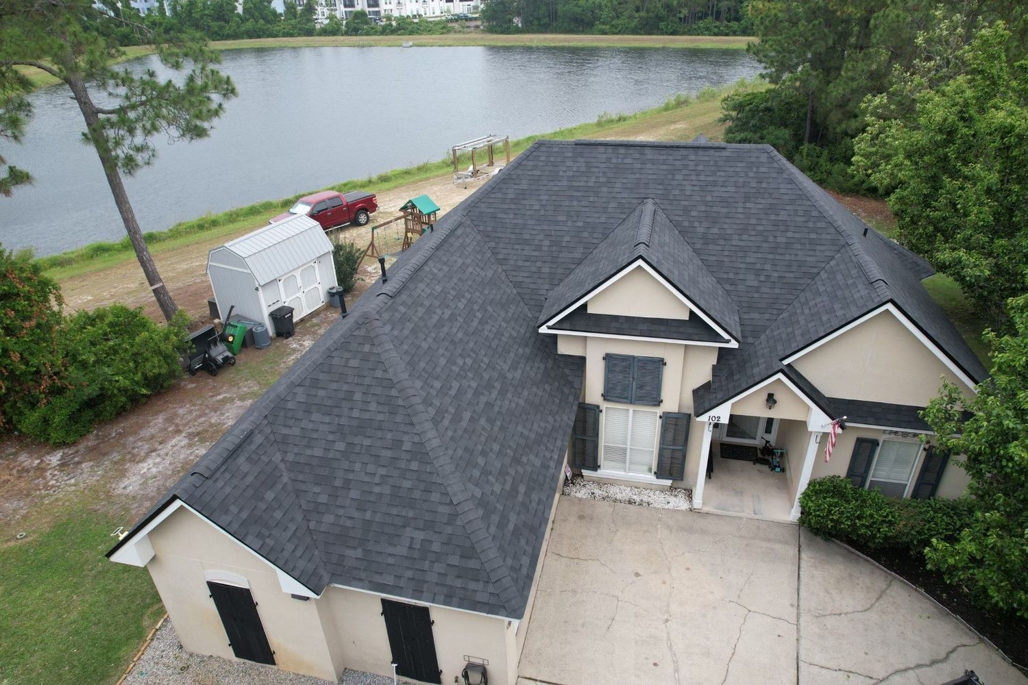 Aerial view of a house with a dark roof beside a lake and trees, with a driveway and parked vehicles.