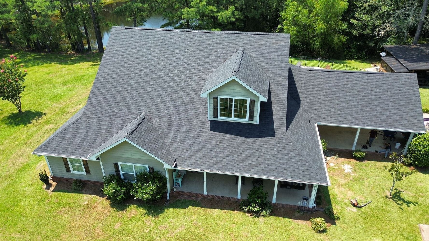 Aerial view of a gray-roofed house with a front porch, dormer windows, and surrounding green trees.