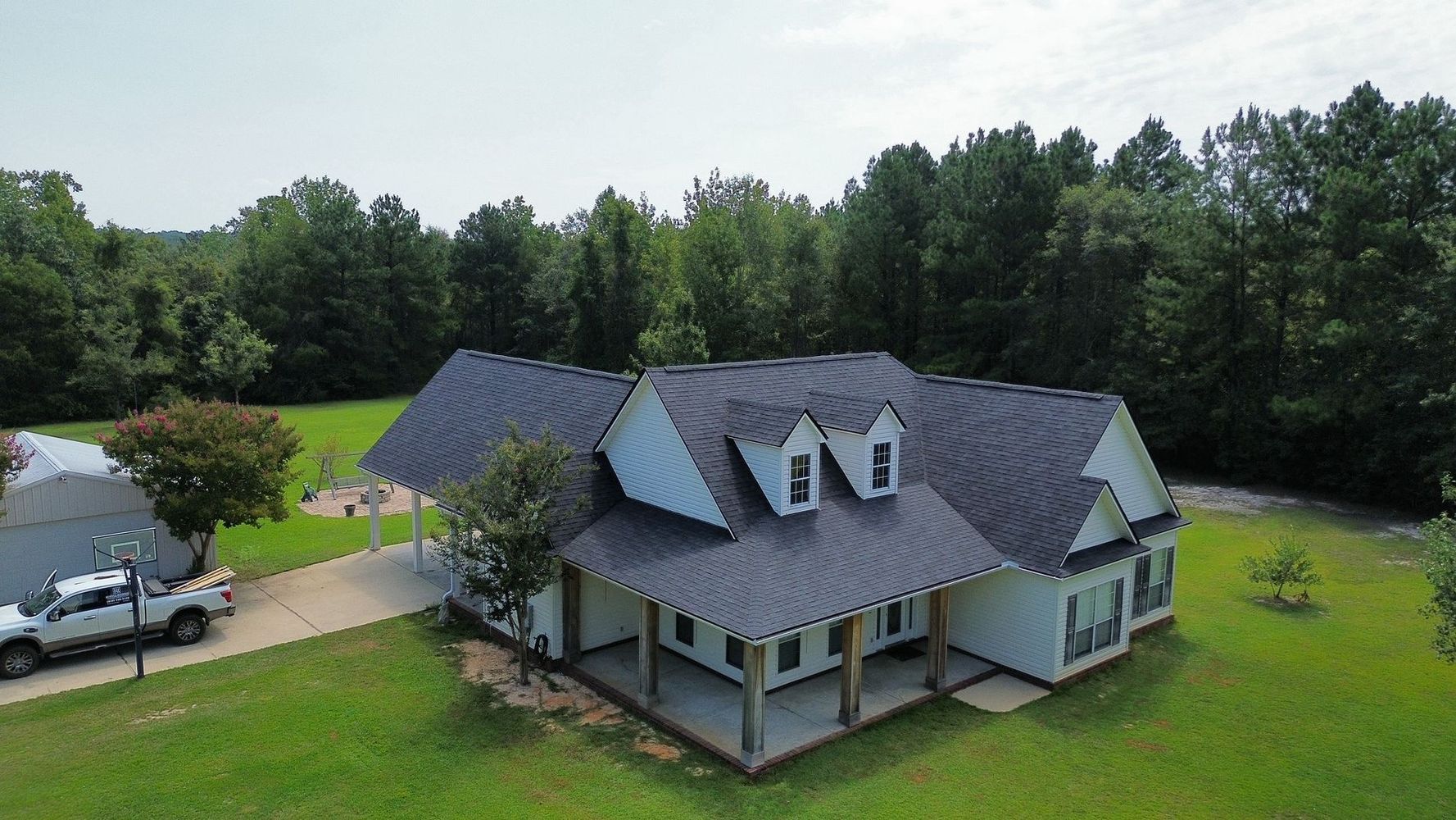 Aerial view of a gray-roofed house with a porch and car, surrounded by green lawn and trees
