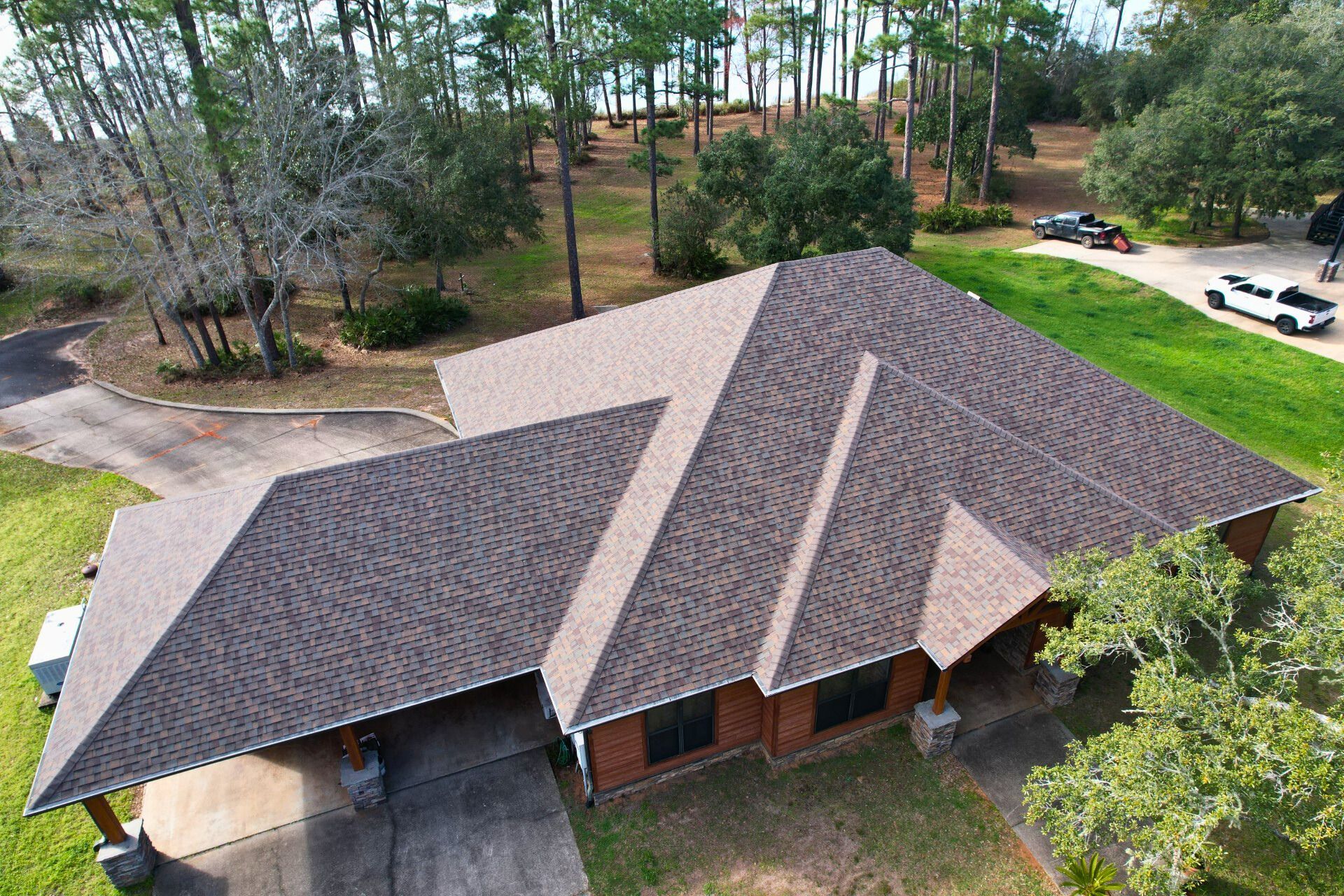 Aerial view of a residential brick house with a dark shingled roof, surrounded by trees and a paved driveway.