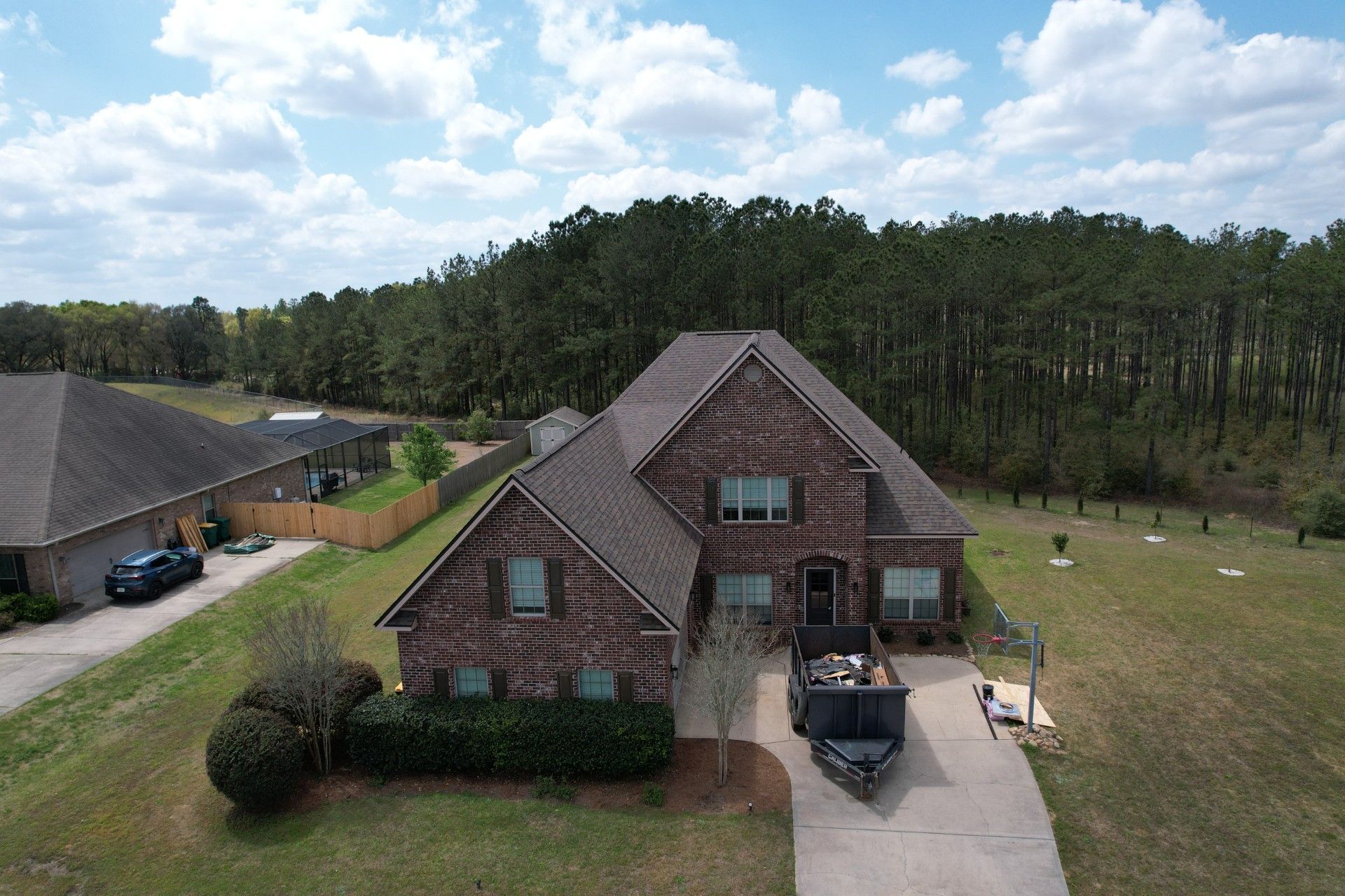Aerial view of a brick house with a driveway, parked SUV, and wooded backyard under clear skies