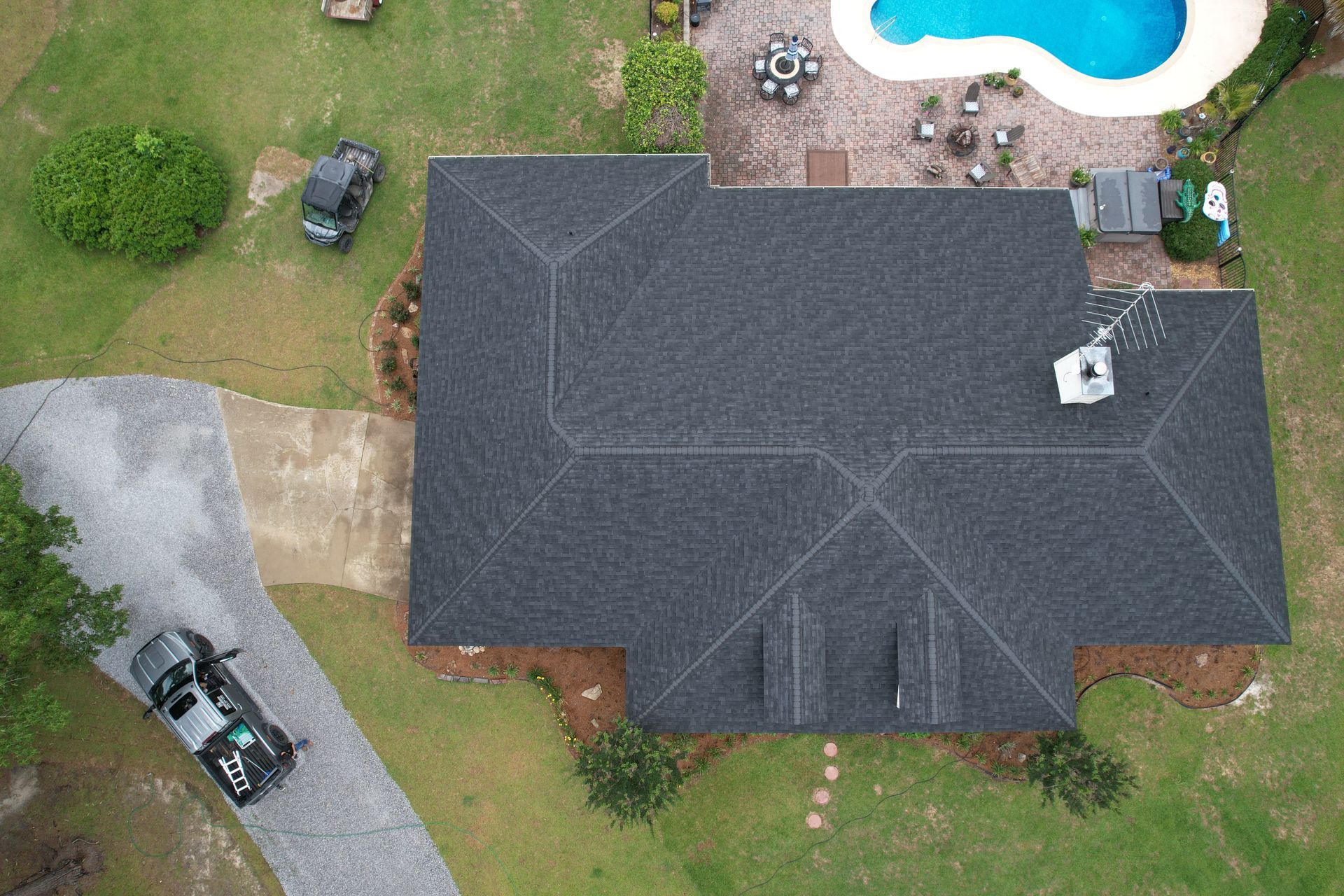 Aerial view of a dark gray roof beside a backyard pool, patio, and driveway with parked vehicles.