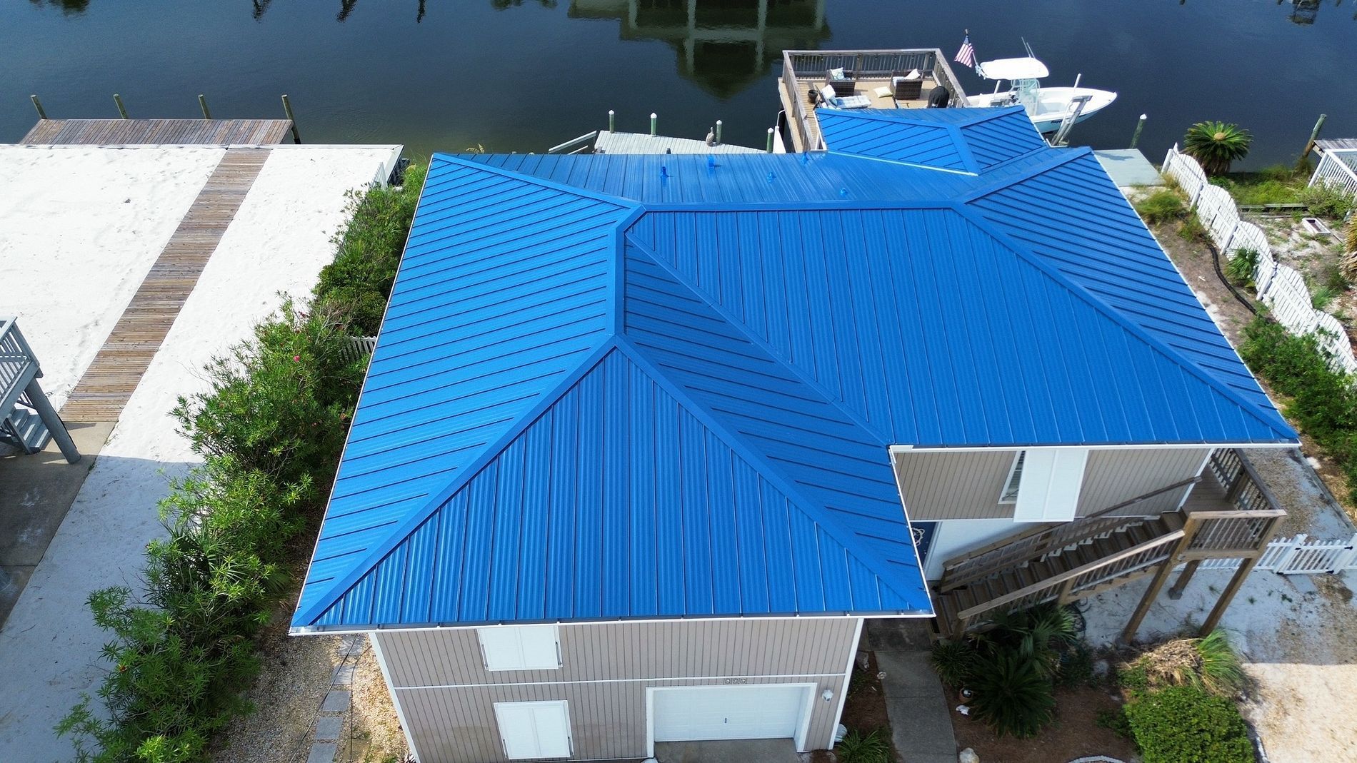Aerial view of a house with a bright blue metal roof beside a canal and docked boats.