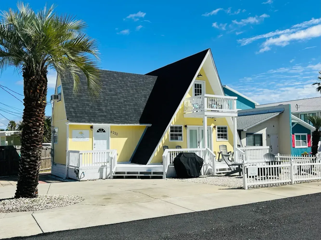 Yellow beach house with black roof, white porch, and palm trees under a bright blue sky.
