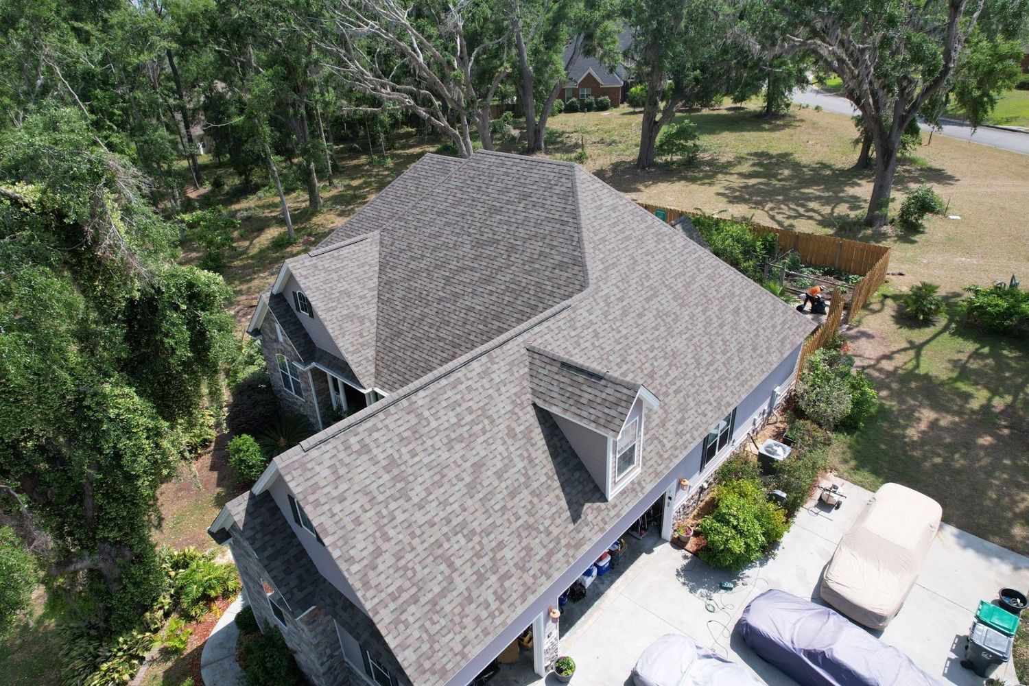 Aerial view of a gray-roofed house surrounded by trees, with vehicles parked beside it.