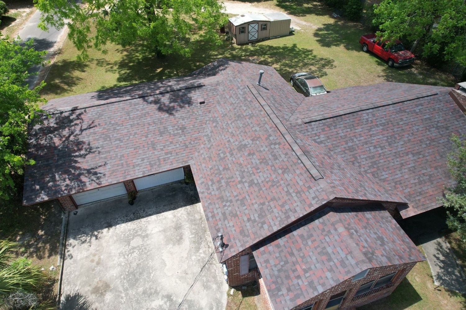 Aerial view of a house with a dark shingled roof, attached garage, and surrounding trees and yard.