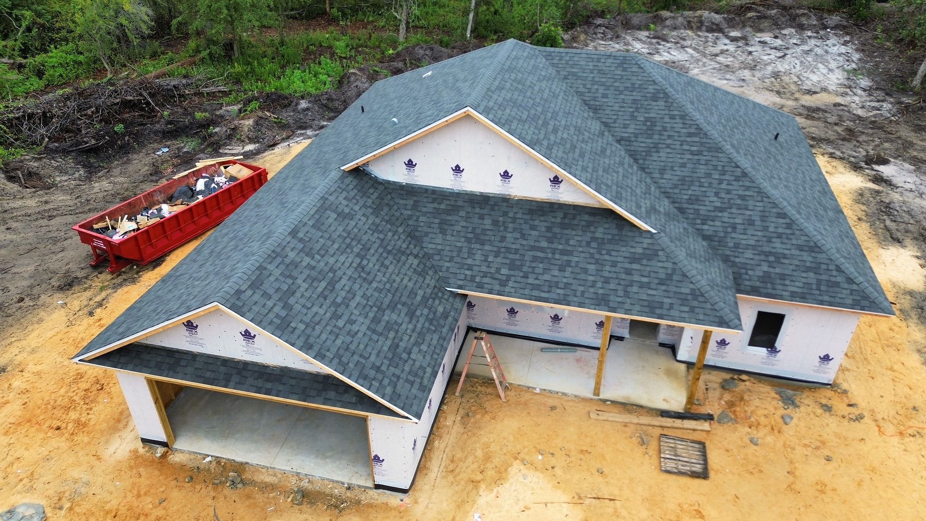 Aerial view of a new gray-roofed house under construction beside a red dumpster on sandy ground.