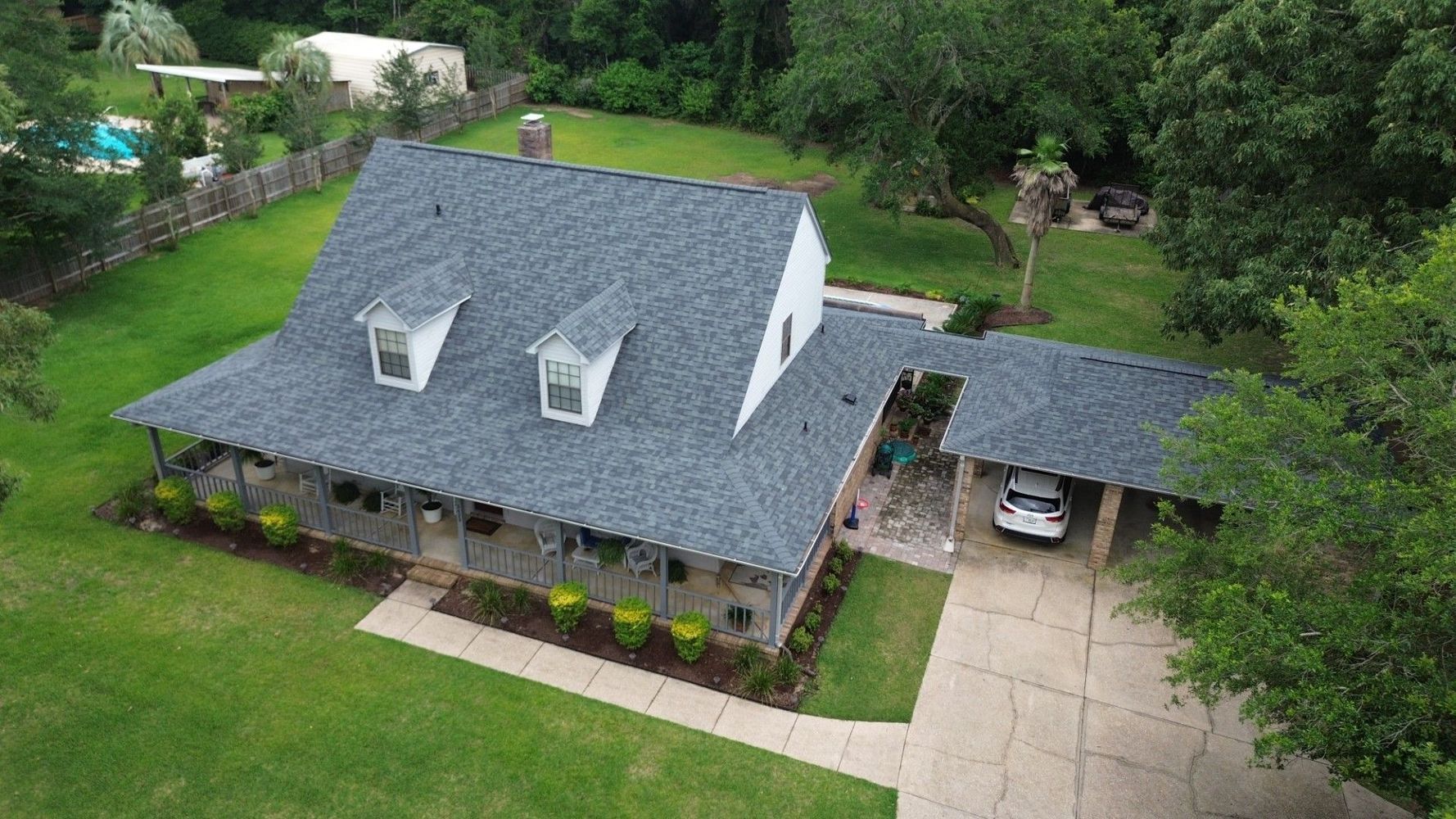 Aerial view of a gray-roofed house with a detached carport, surrounded by green lawn and trees.