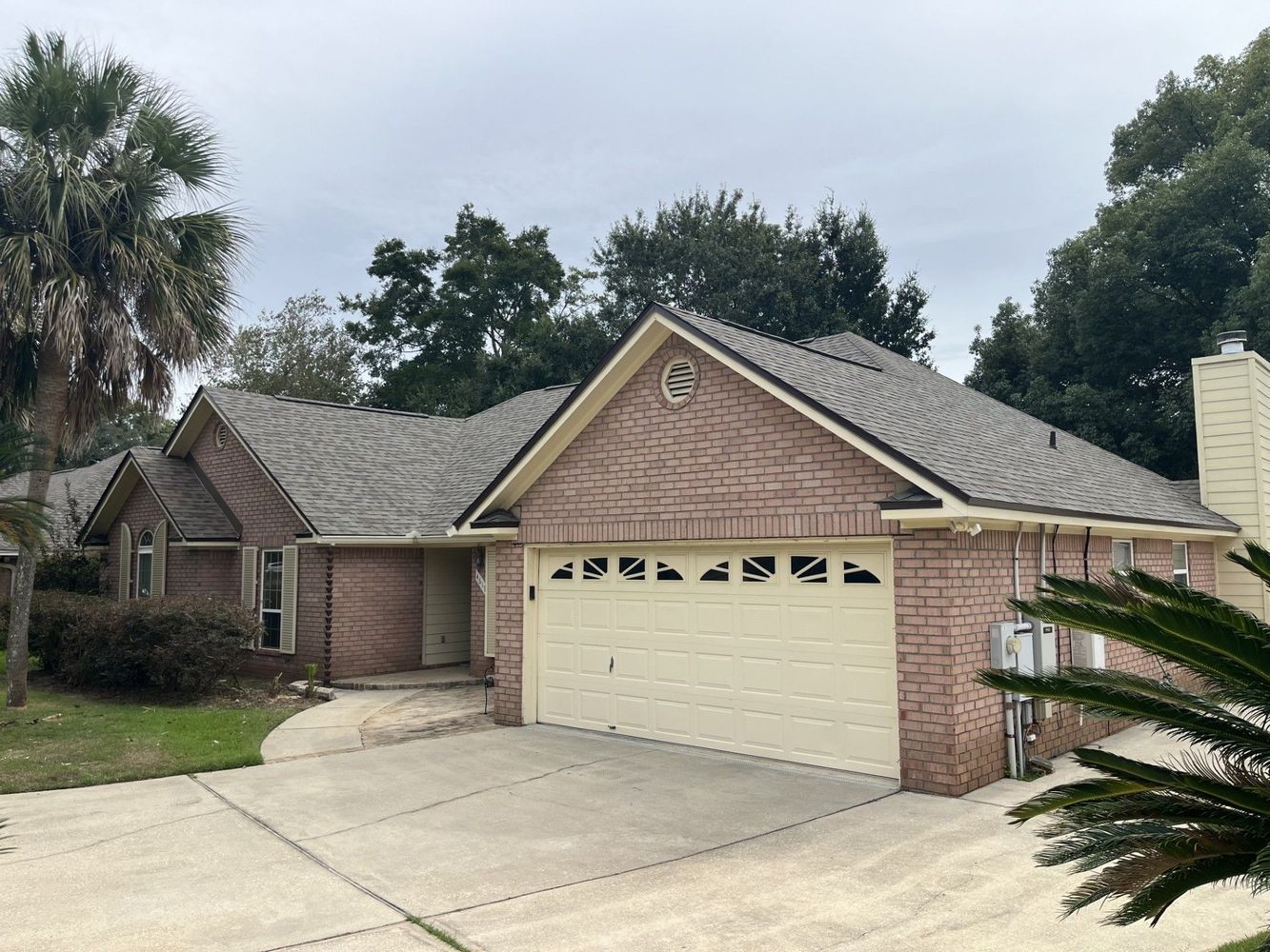 Suburban brick house with a cream garage door, driveway, and trees under a cloudy sky