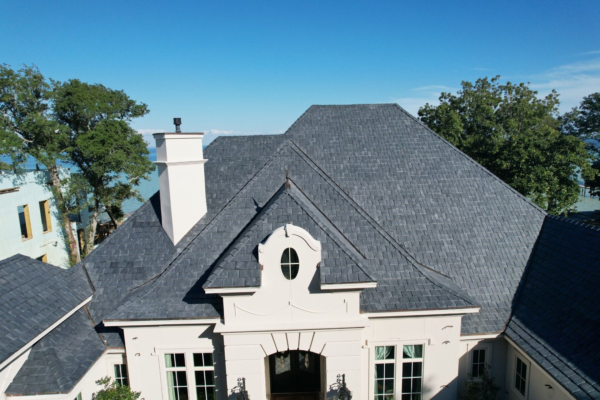 Aerial view of a white house with a dark gray shingled roof and chimney, surrounded by trees