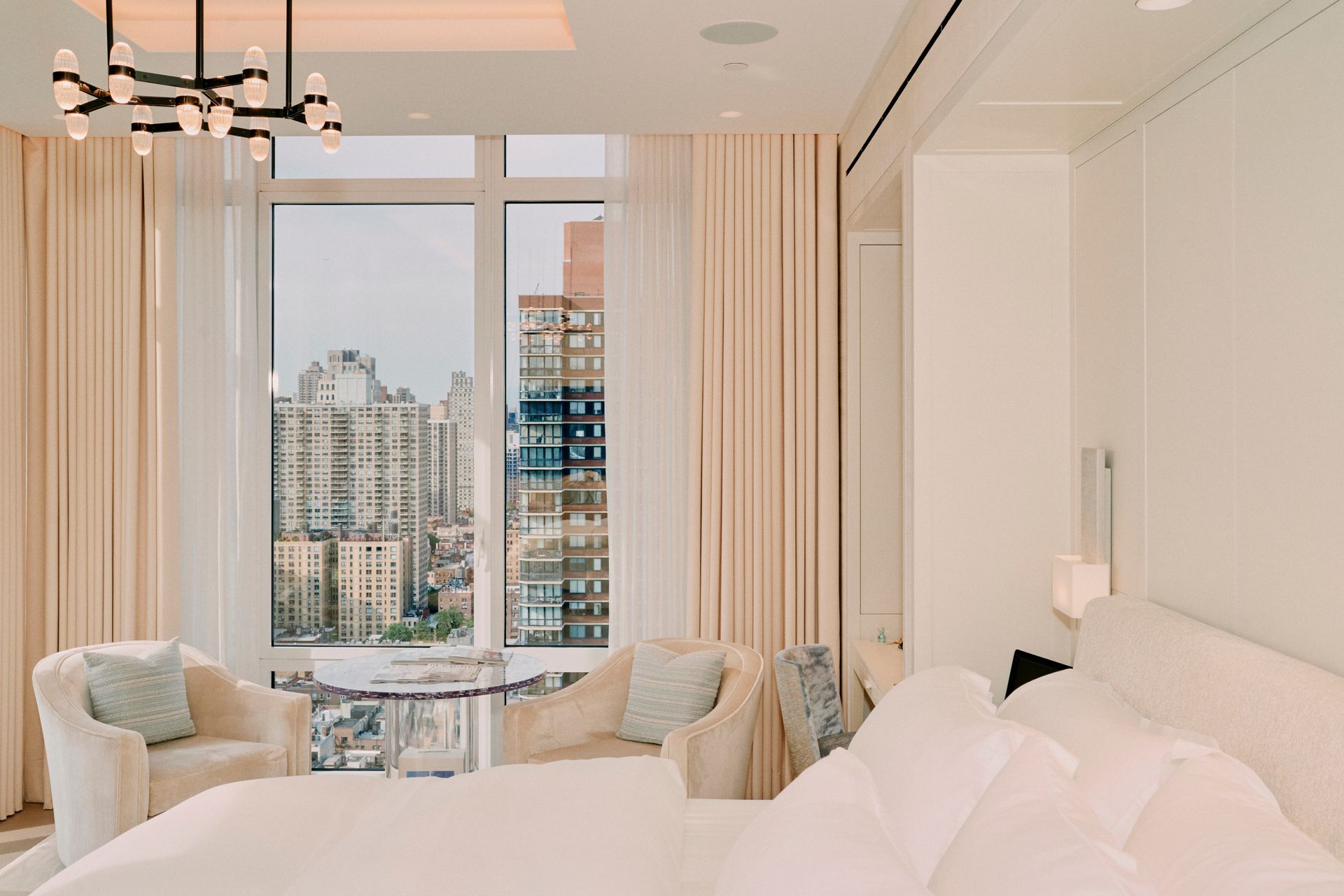Elegant bedroom with city view; cream-colored drapes, bed, and chairs; glass table and chandelier.