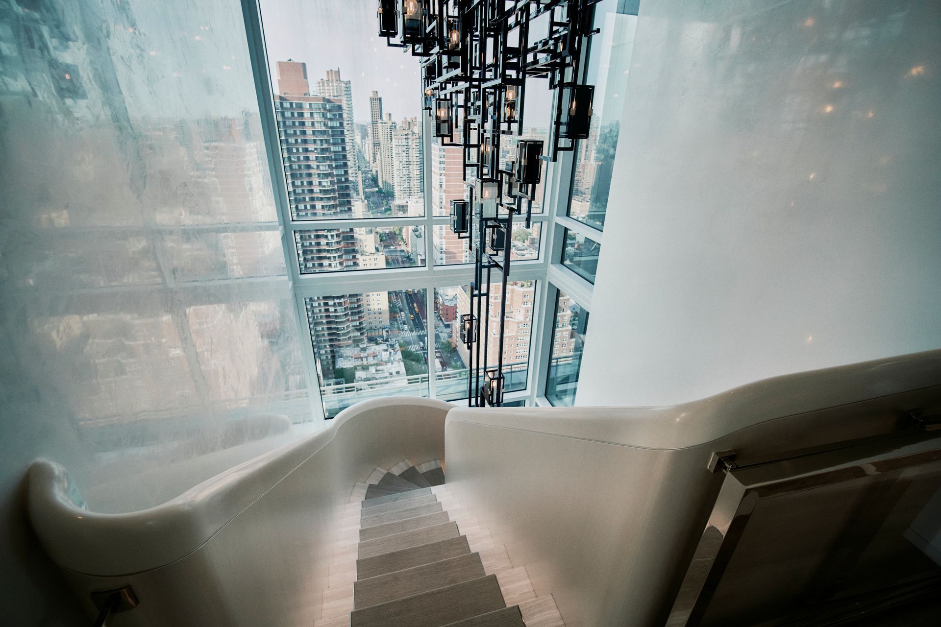 Staircase with modern, white design, leading down with a city view through large windows and a chandelier.