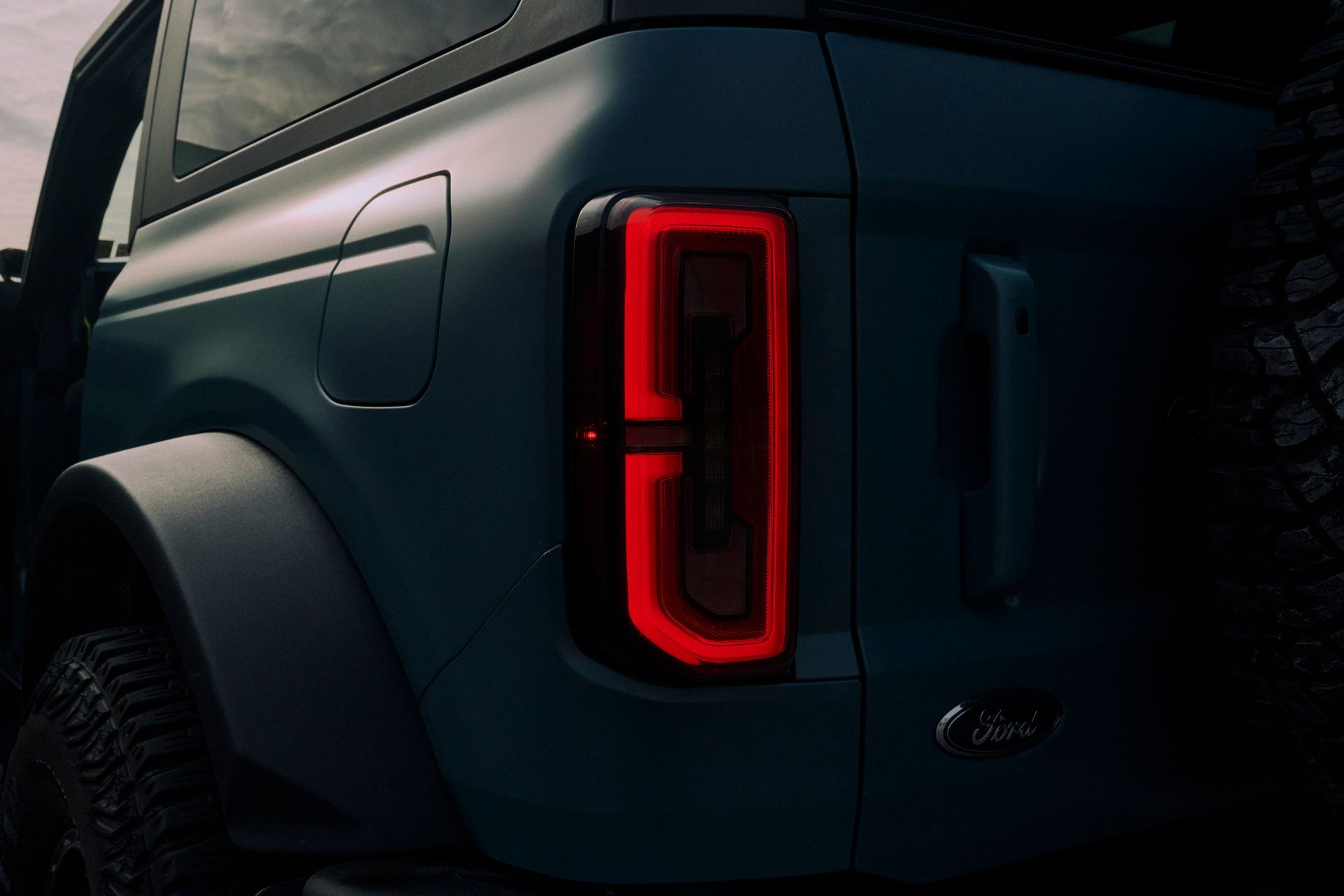Rear view of a teal Ford Bronco, showing a lit red taillight and spare tire.