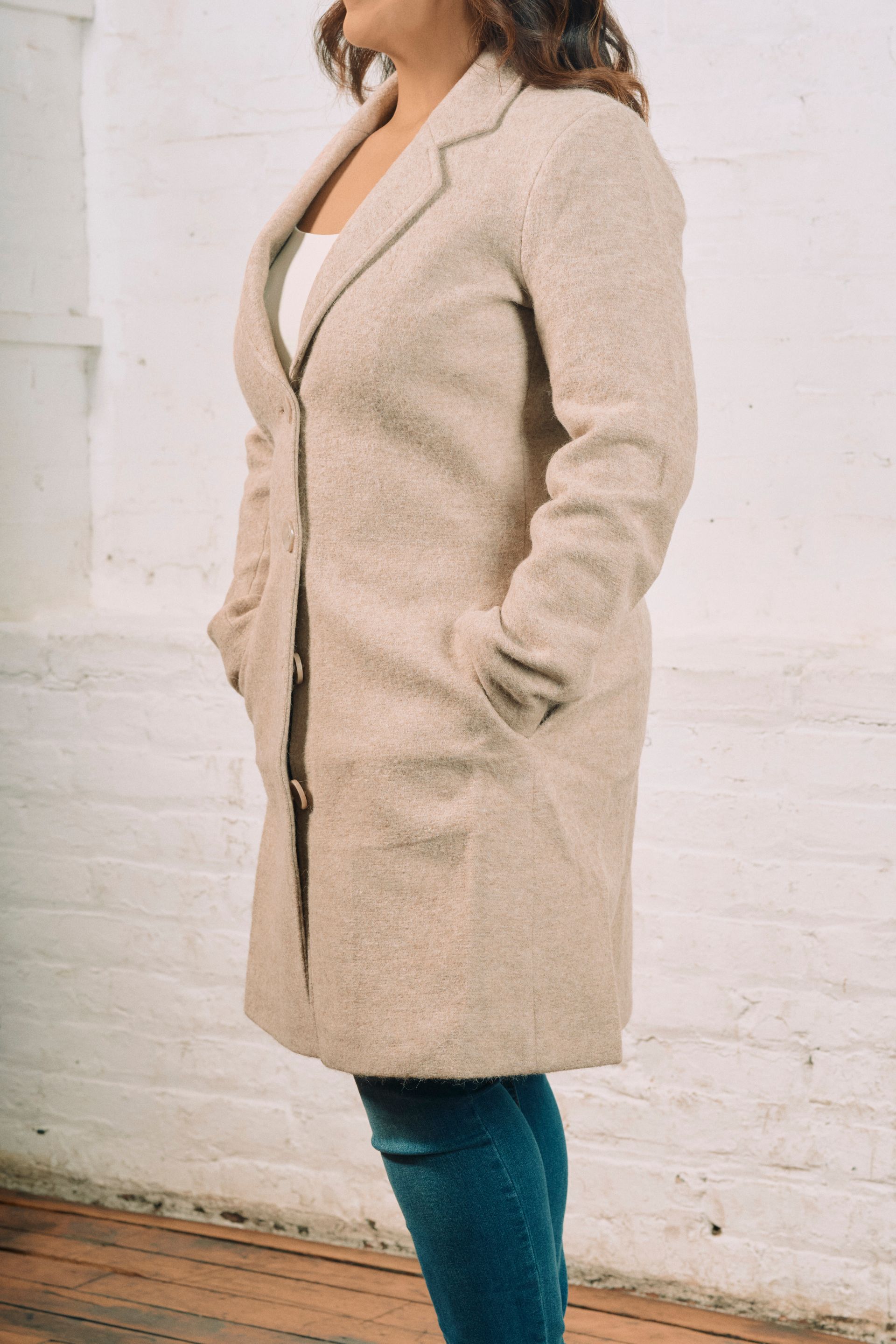A woman is standing in front of a white brick wall wearing a beige coat.