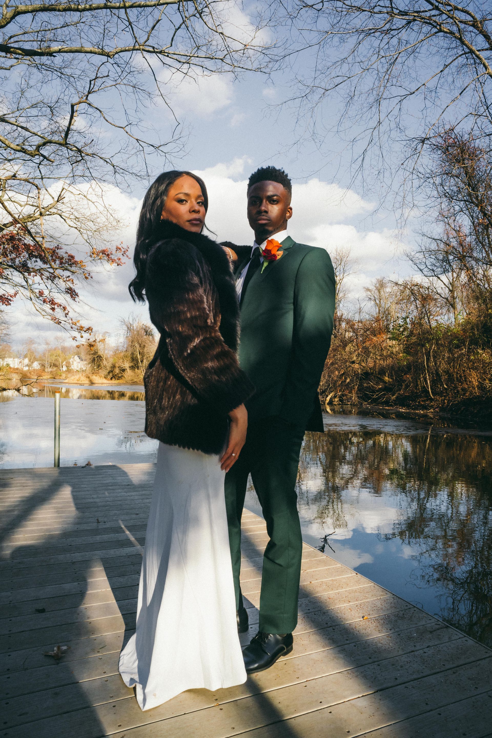 Couple on a wooden dock with lake in the background; the woman in a fur coat, the man in a green suit.