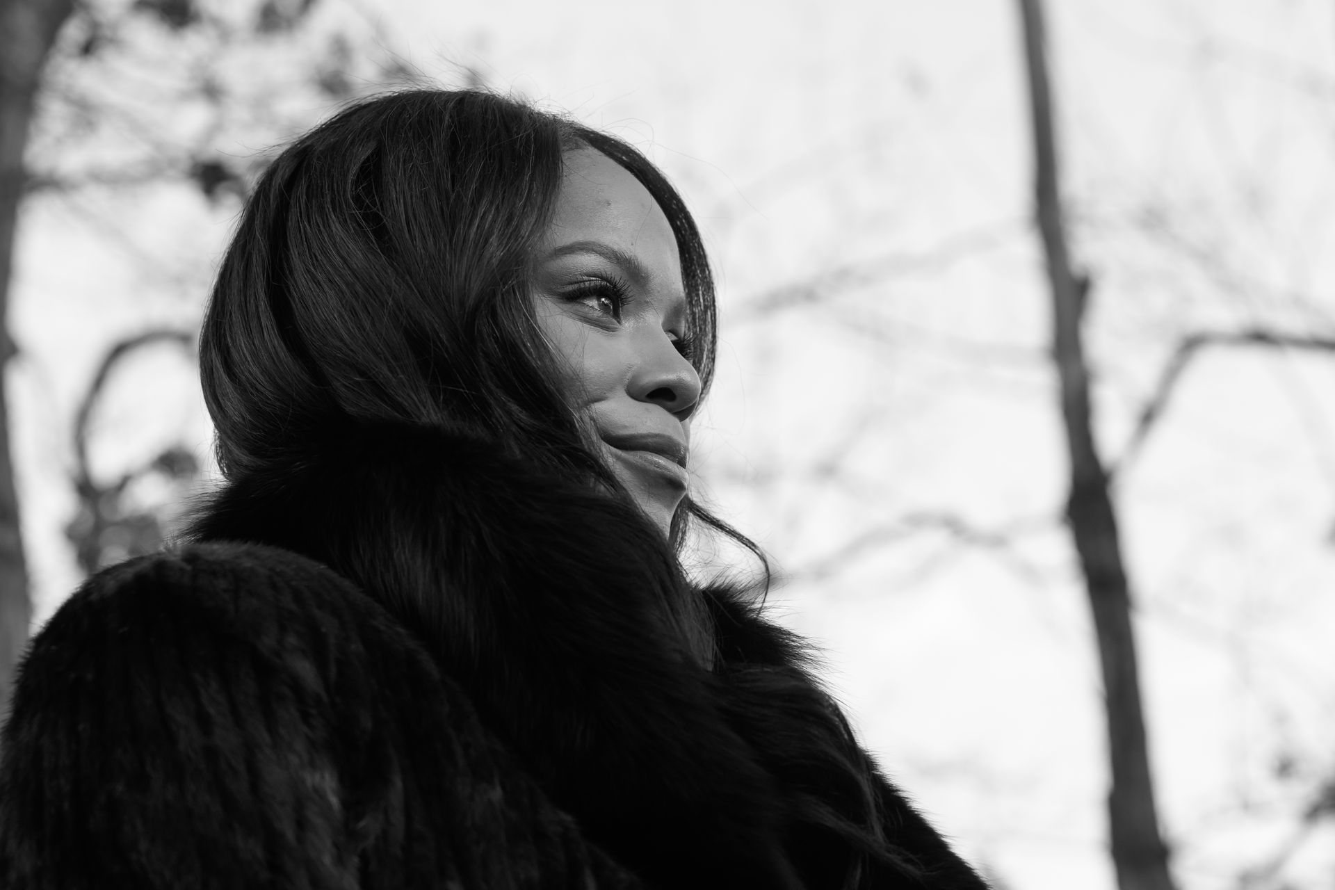Woman in dark fur coat looks off to the side, outdoors with blurred trees in background.