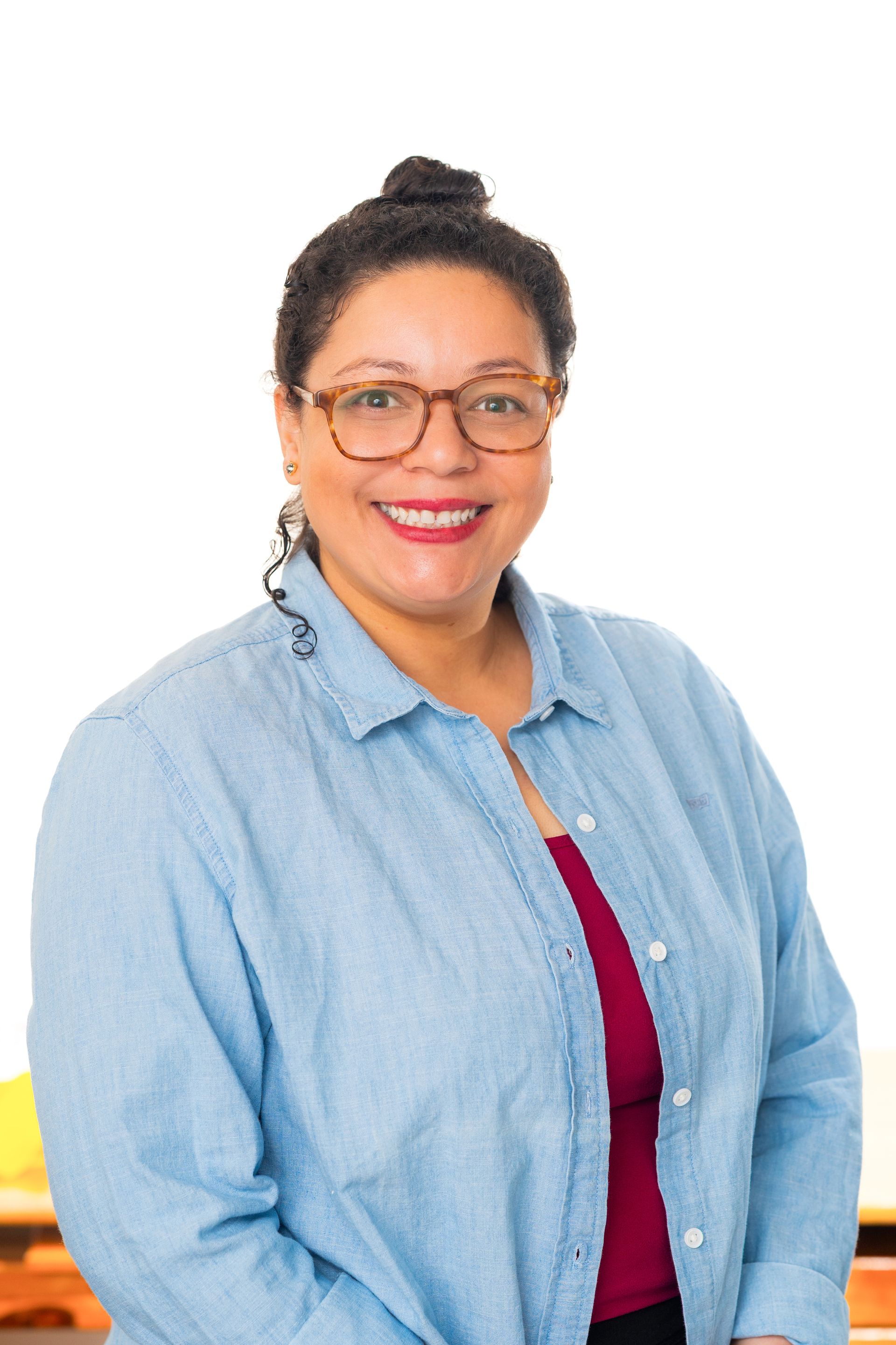 Woman with glasses smiles, wearing denim shirt over a red top. White background.