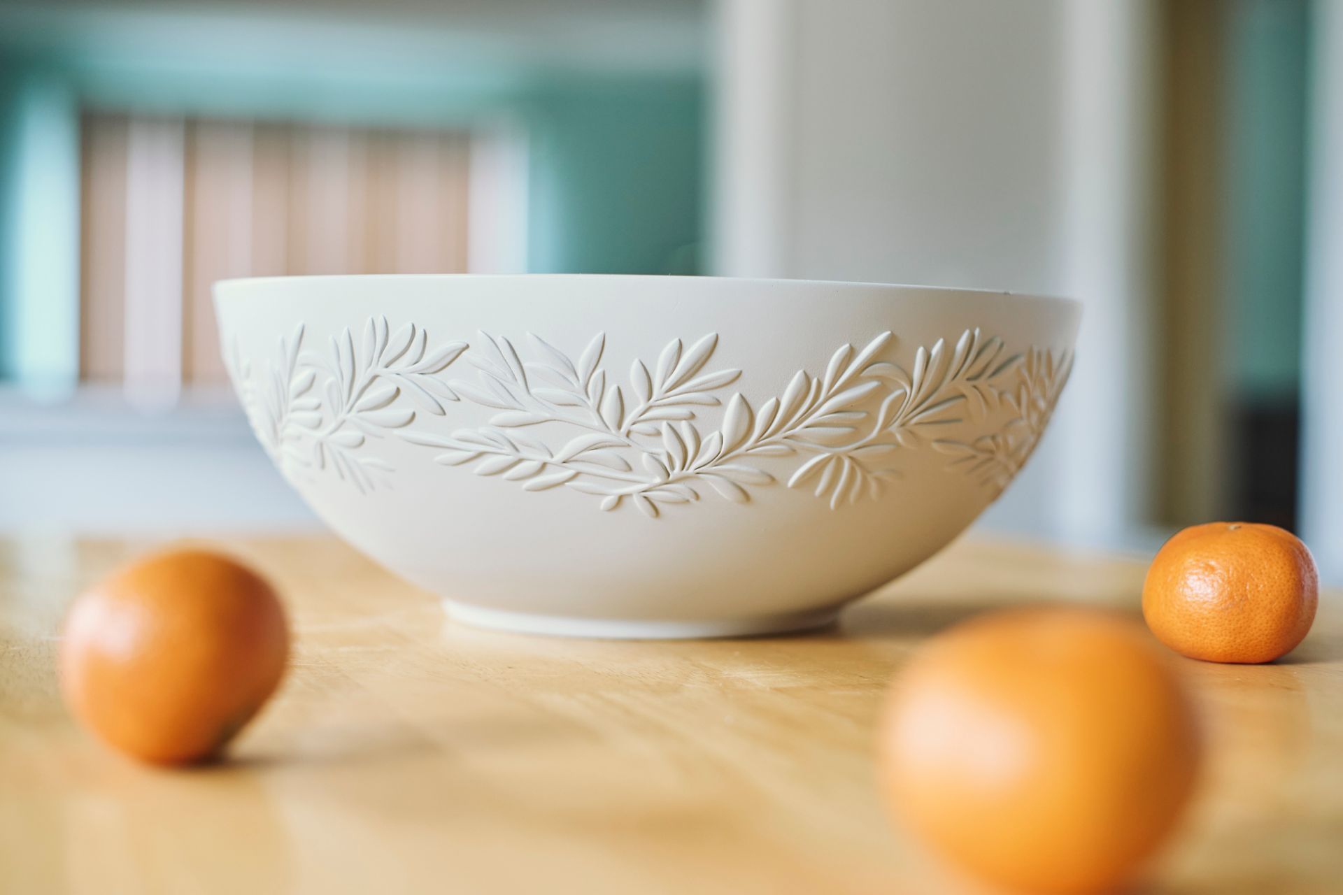 White bowl with leaf design, sits on wood table with three oranges.