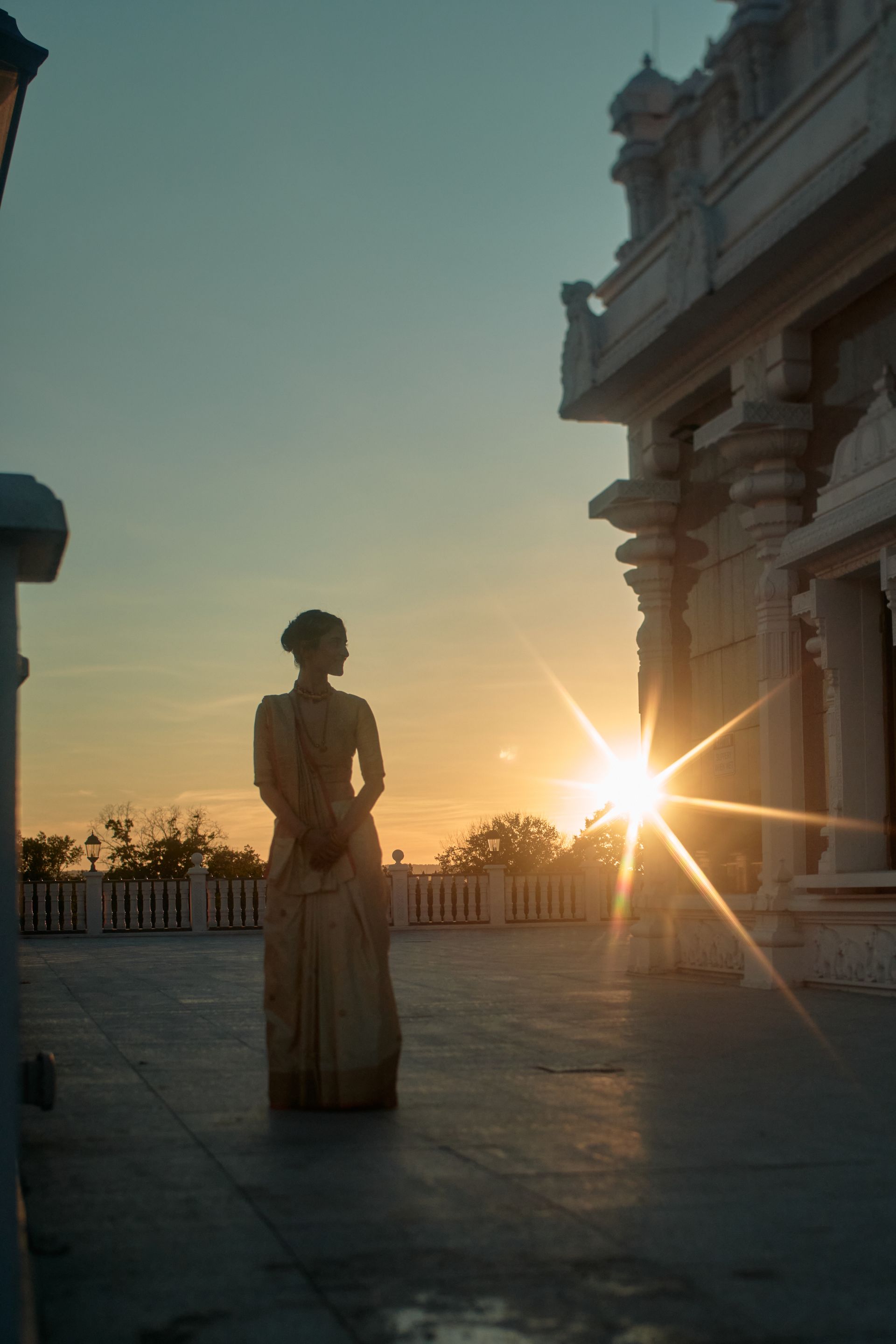 Silhouetted statue at sunset beside a historic building, with sun flaring near the horizon