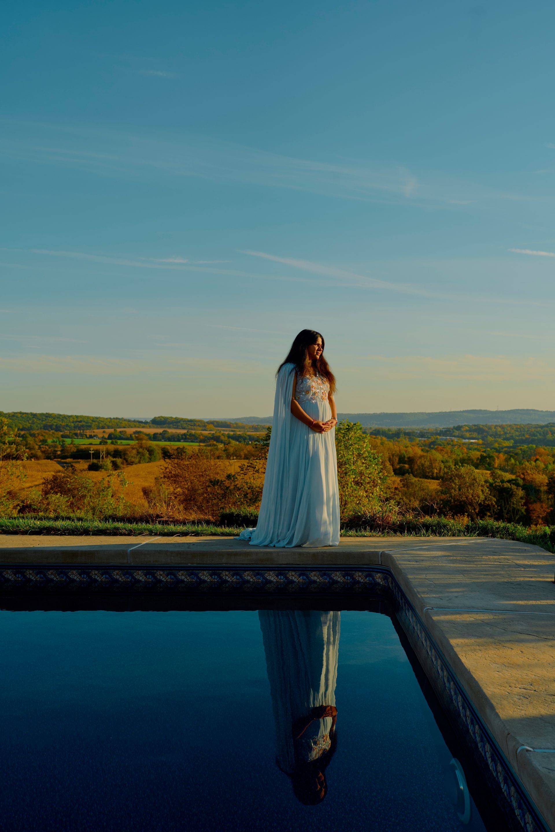Pregnant person in white gown stands by pool overlooking autumn landscape.