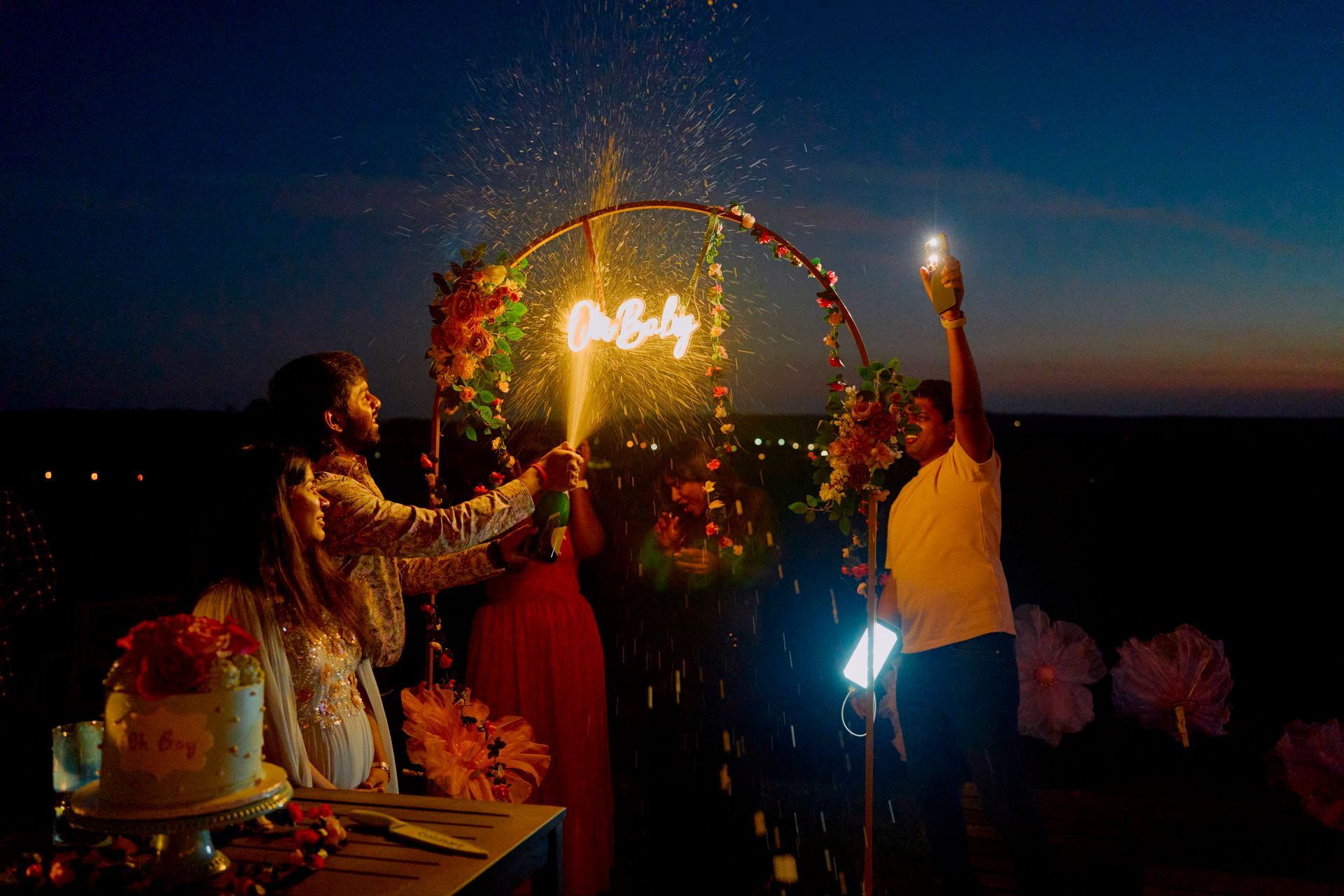People lighting sparklers around a floral hoop with illuminated initials at a night event. Cake on table.
