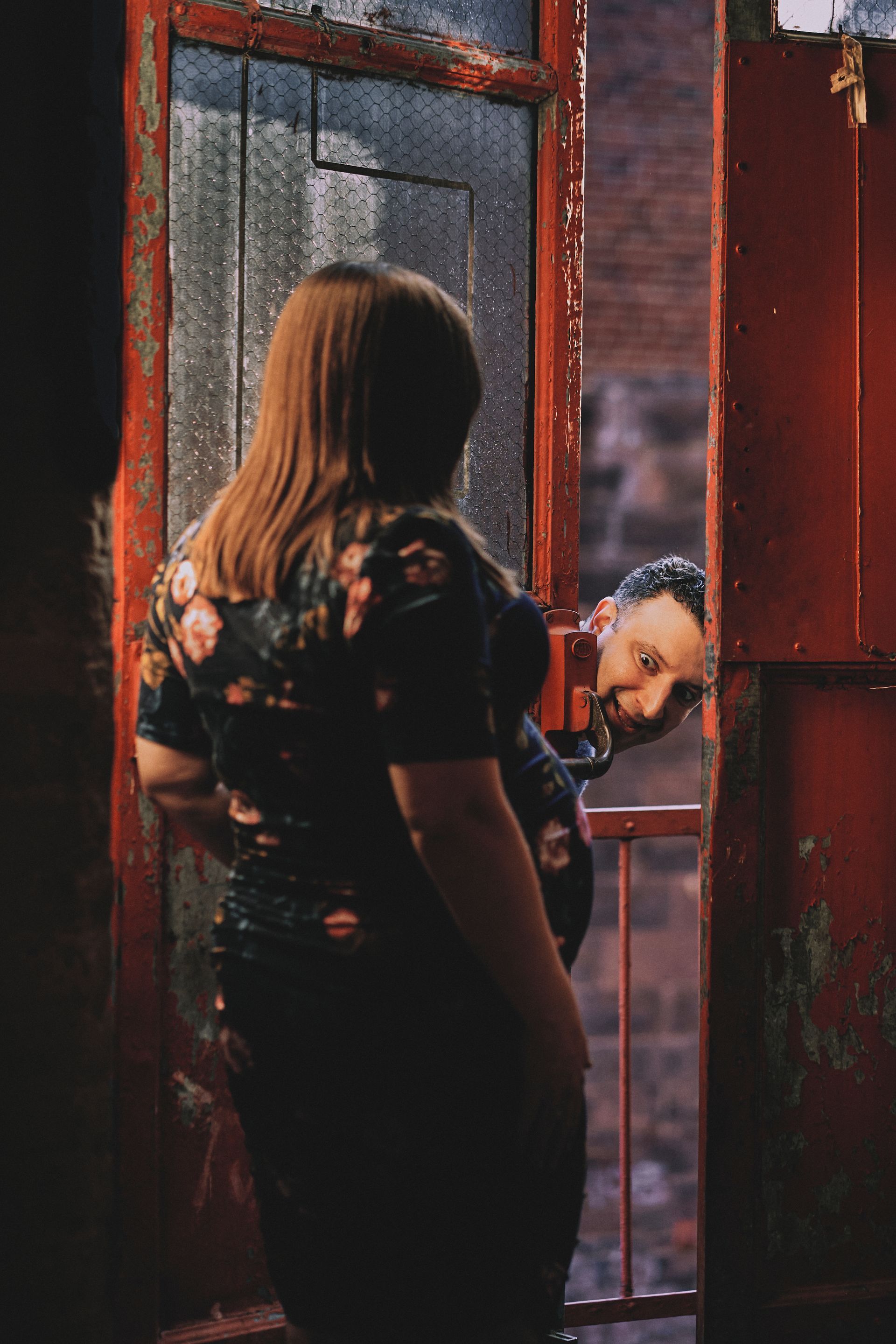 Woman in floral top looks out window, another person peeks in from the side. Red door frame, brick background.
