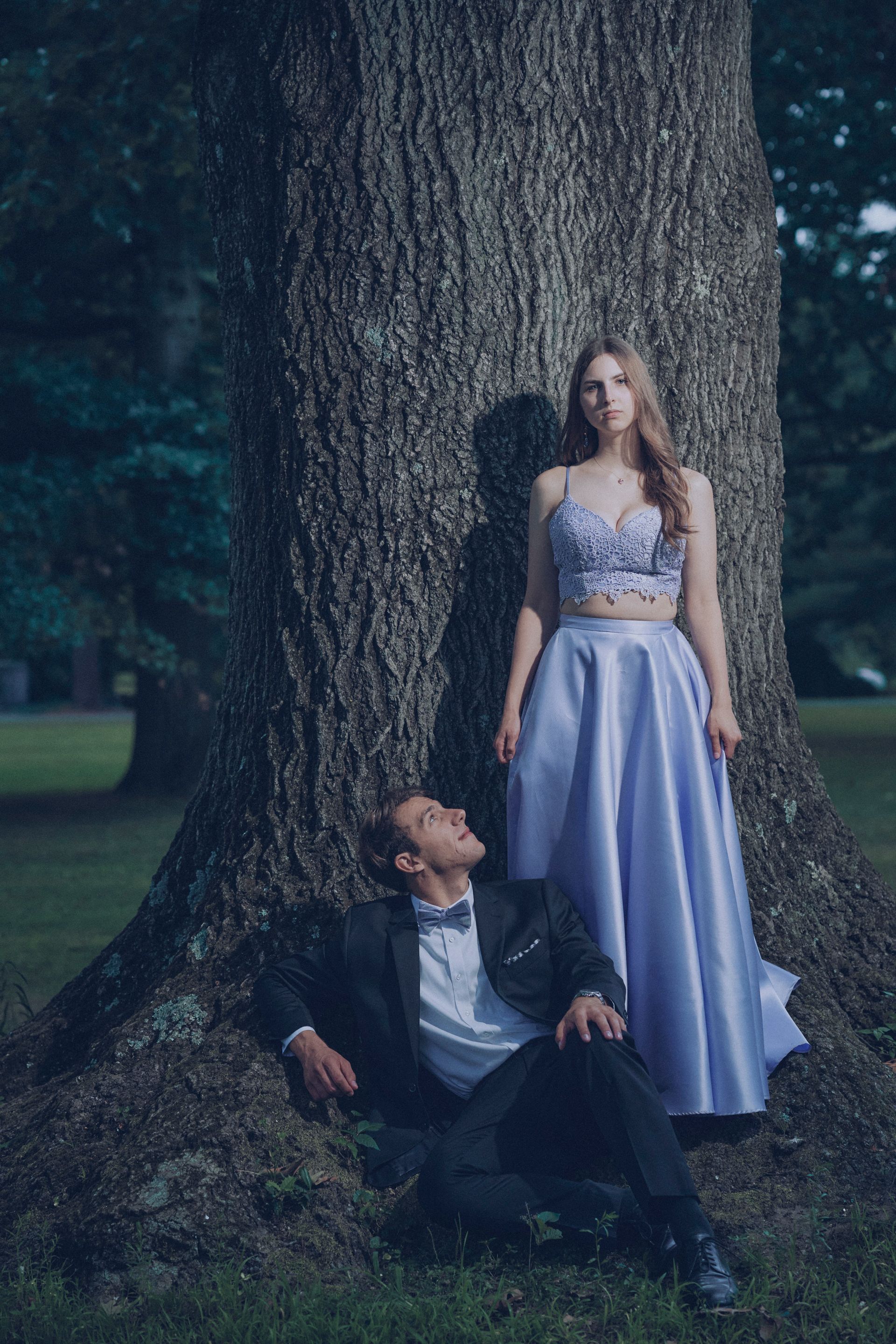 A man and a woman are standing next to a tree . the woman is wearing a purple dress.