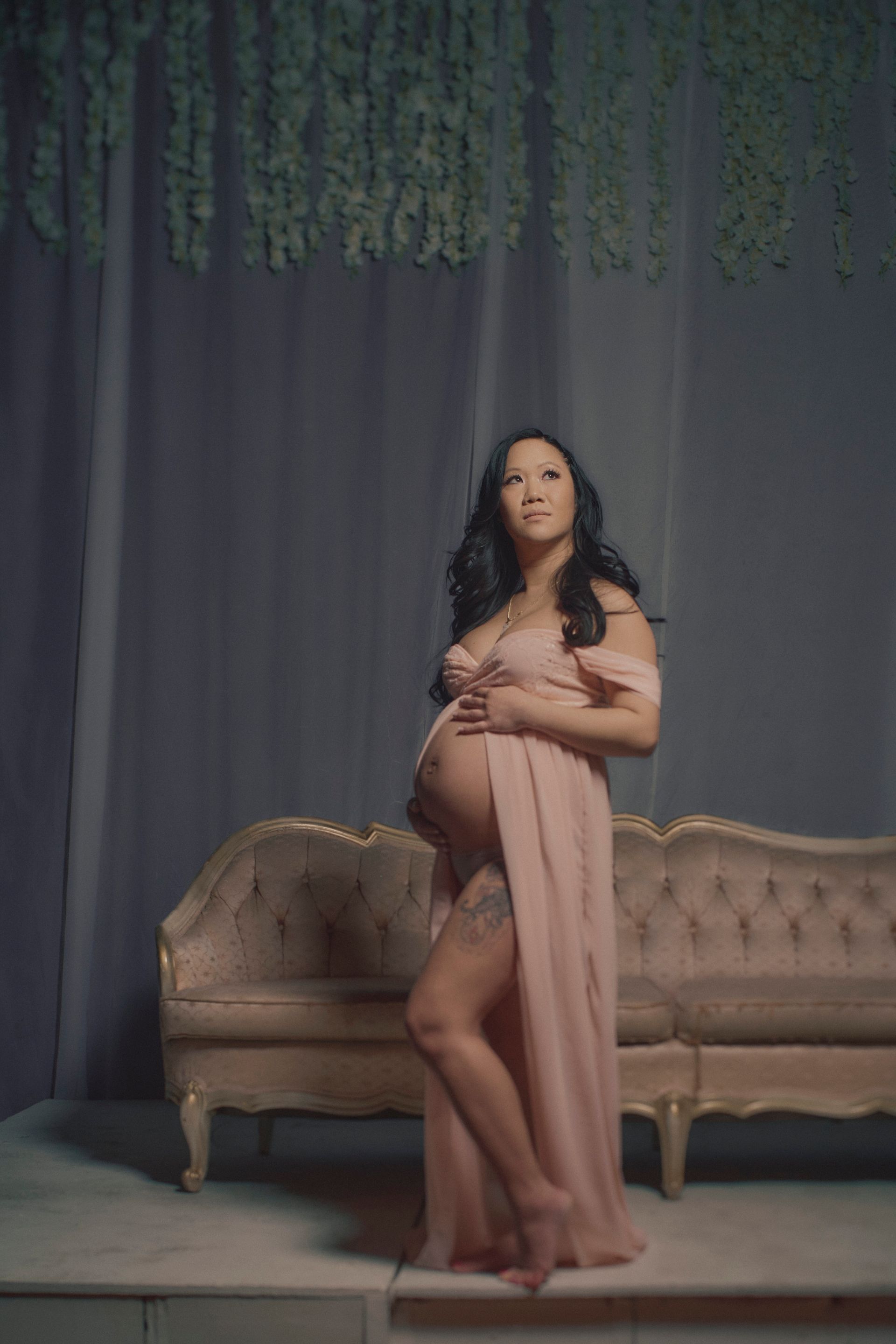 A pregnant woman in a pink dress is standing in front of a couch.