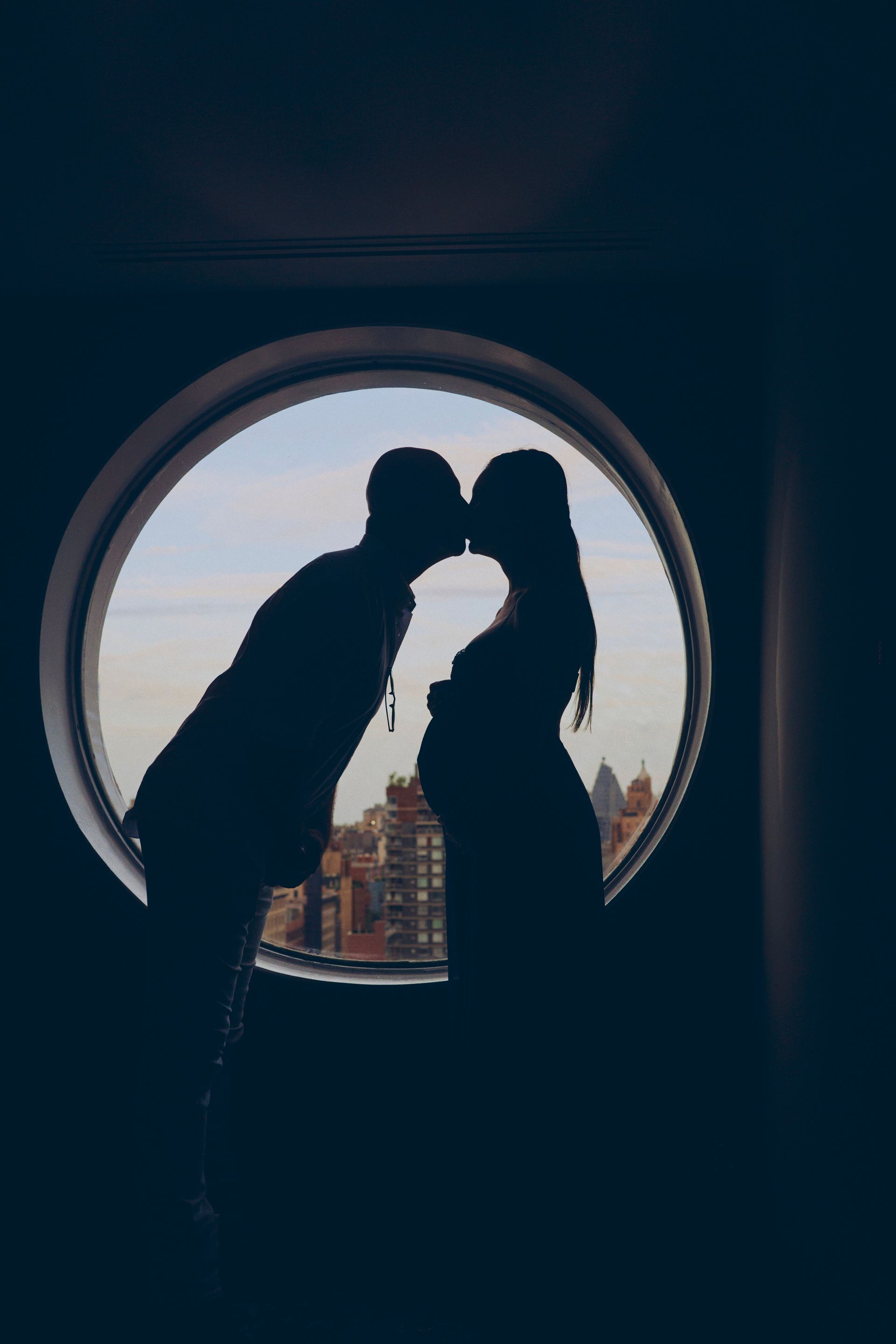 Silhouette of a couple kissing in front of a round window, the woman visibly pregnant with city view.
