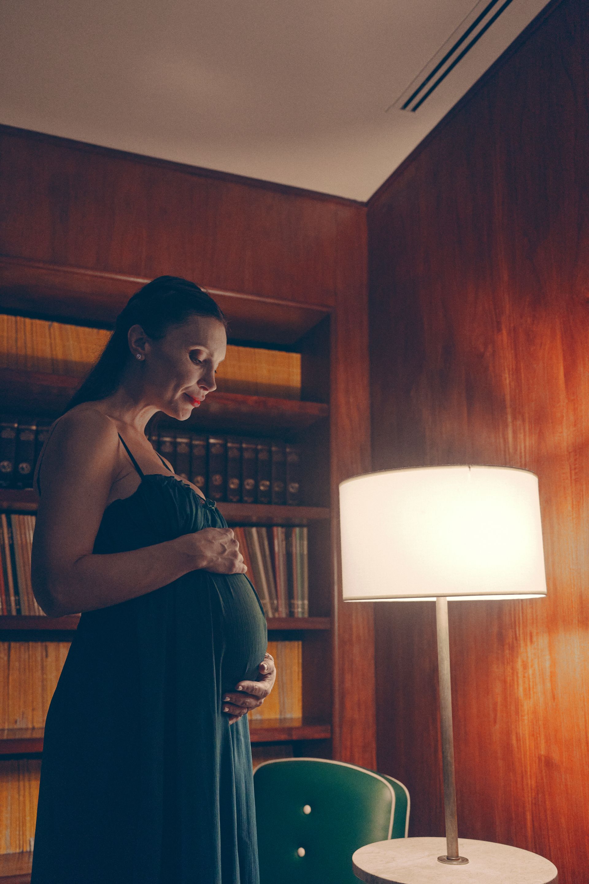 Pregnant person holding belly, standing in a library. Dark dress, wooden bookshelves, lamp lit.