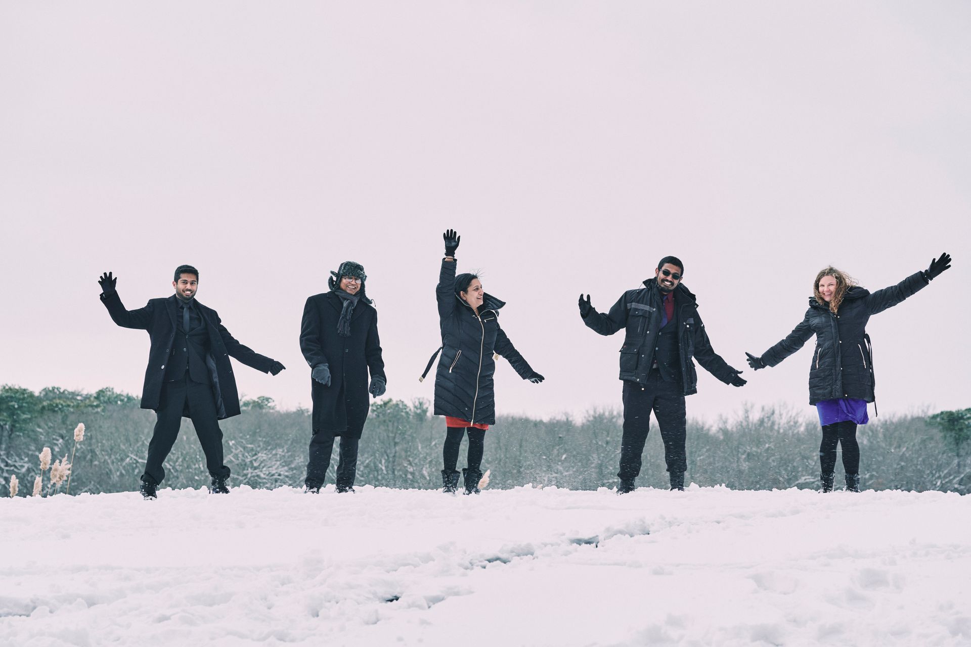 Five people in winter coats jump in a snowy field with arms outstretched.