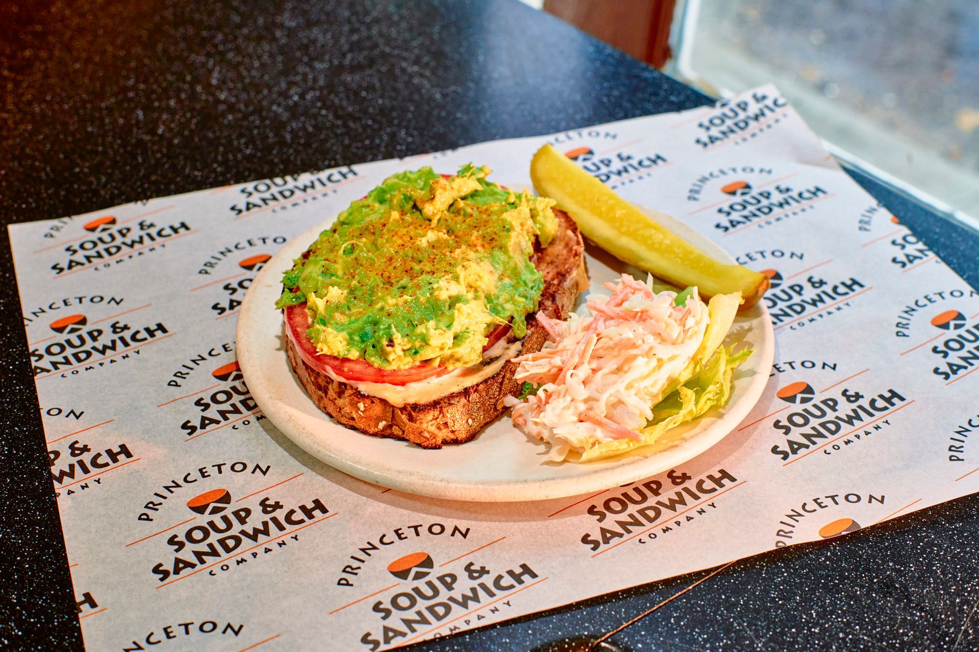 Sandwich with coleslaw, pickle, served on branded paper.