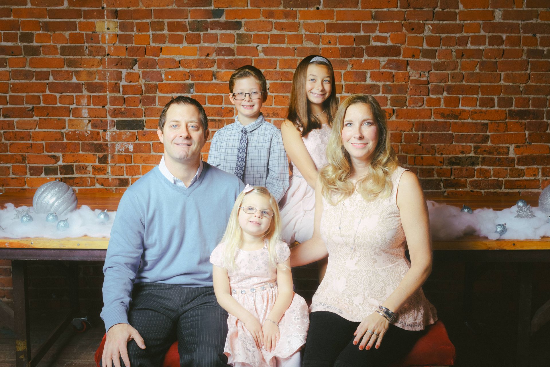 Family portrait in front of a brick wall. Parents and three children smiling, wearing light colors. Snowflakes and decorations.