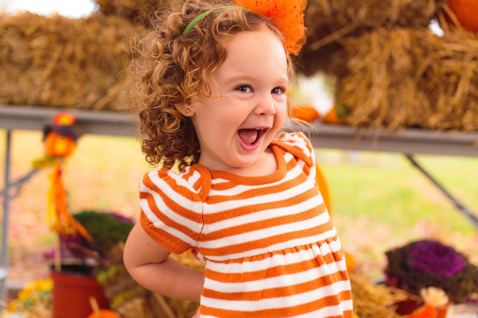 Girl with curly hair, wearing orange and white striped dress, smiles widely, at fall festival.