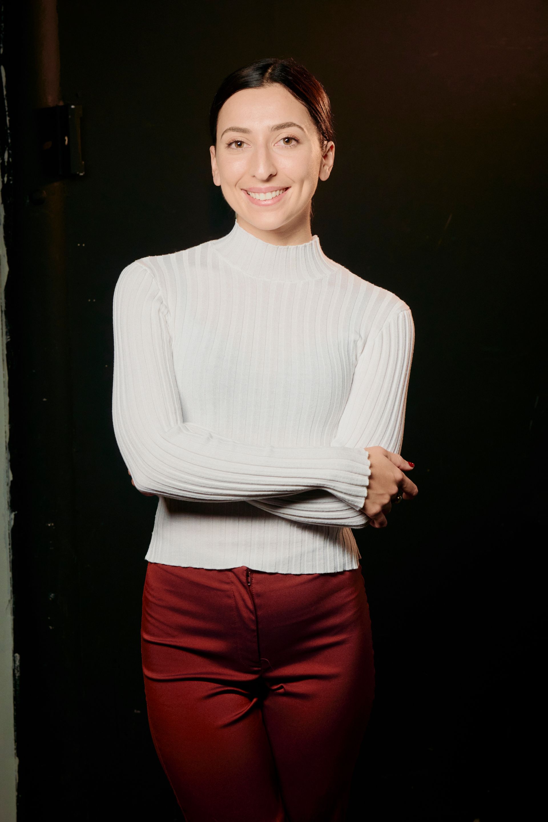 A woman in a white sweater and red pants is standing with her arms crossed.