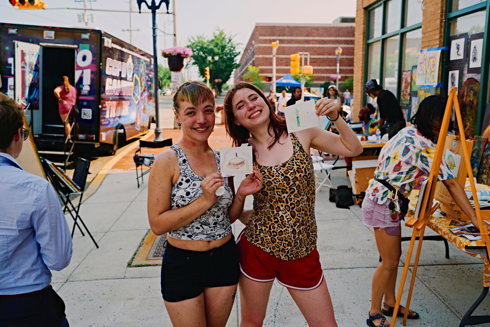 Two smiling people holding a card at an outdoor event with art and a decorated trailer.