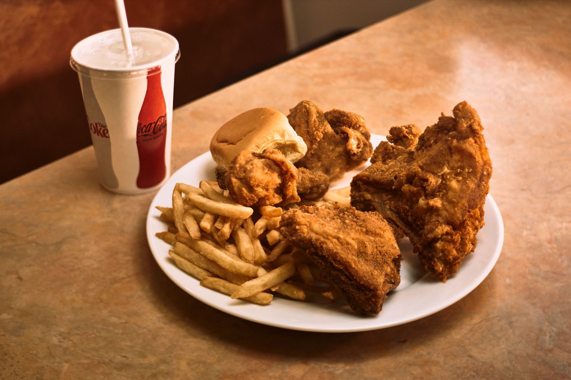 Fried chicken meal on a plate with fries, a biscuit, and a soft drink in a restaurant setting.