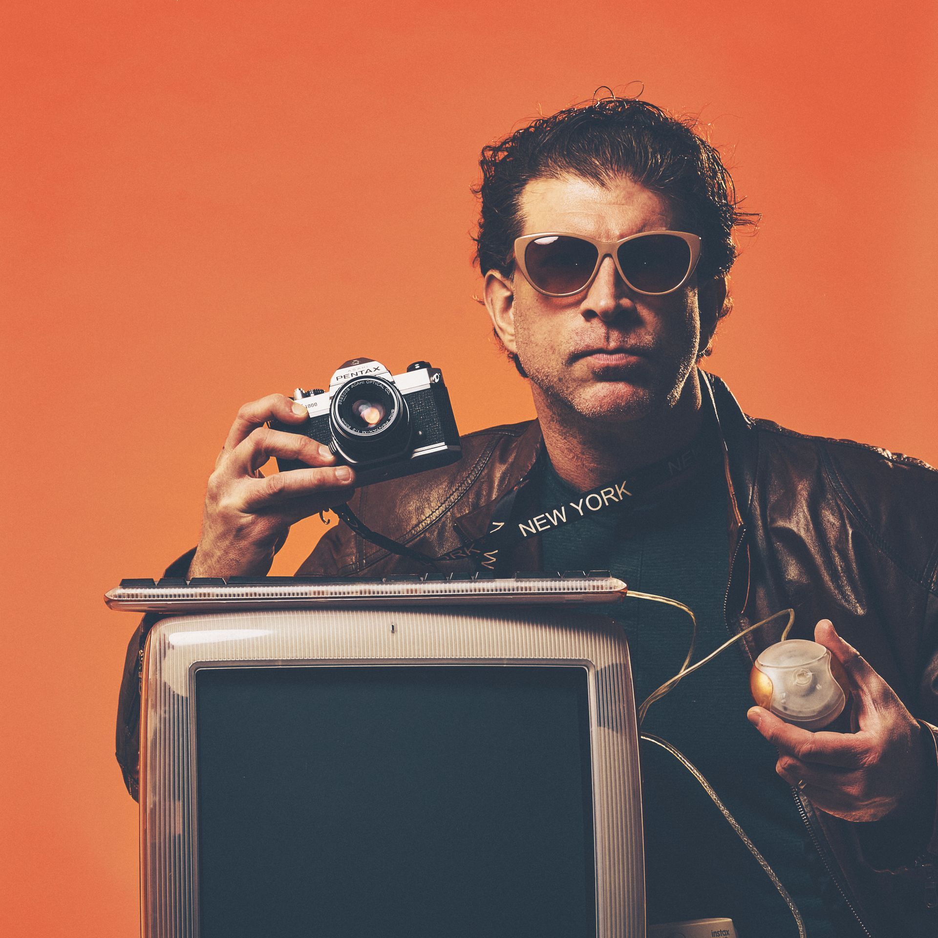 Man in sunglasses holds camera, orange background.