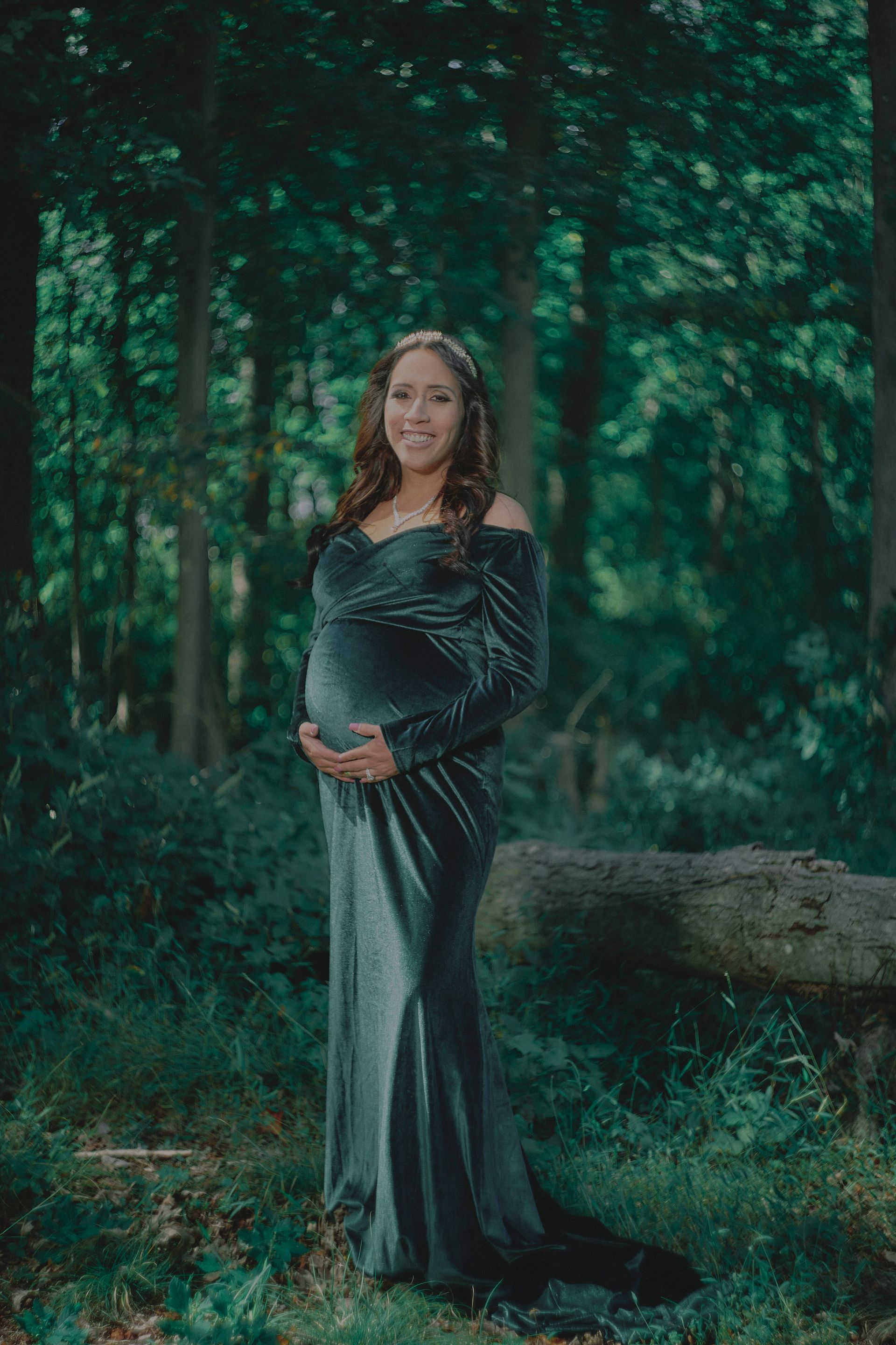 A pregnant woman in a long green dress is standing in the woods holding her belly.