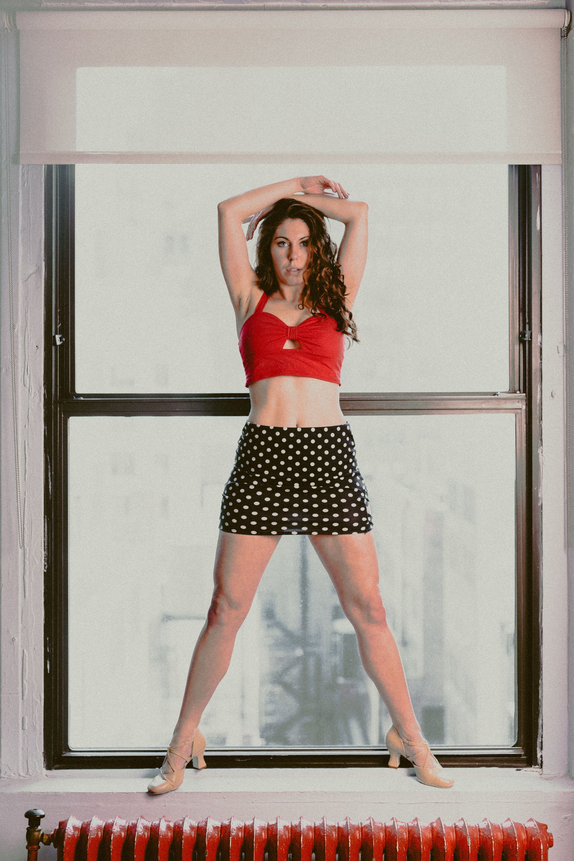 A woman in a red top and polka dot skirt is standing in front of a window.