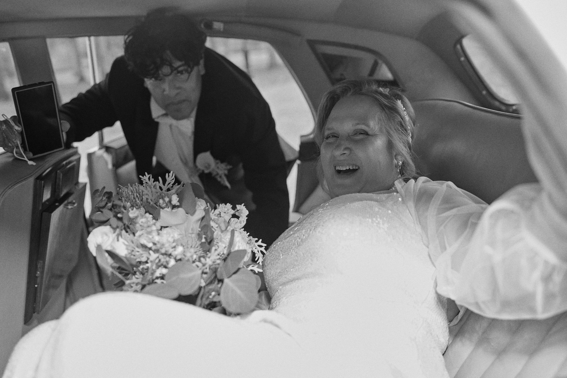 Bride in white dress smiles, bouquet in lap. Groom behind her, holding phone in car.