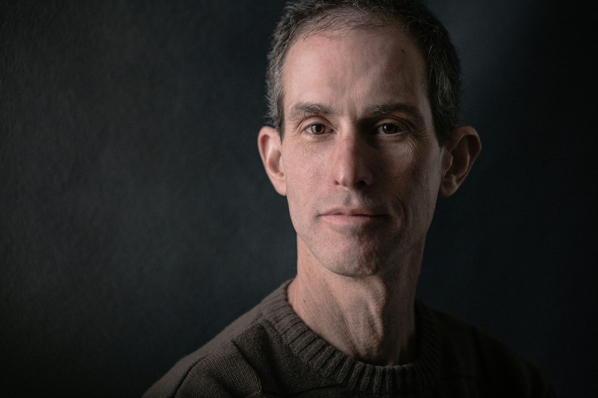 A man in a sweater is looking at the camera with a dark background.