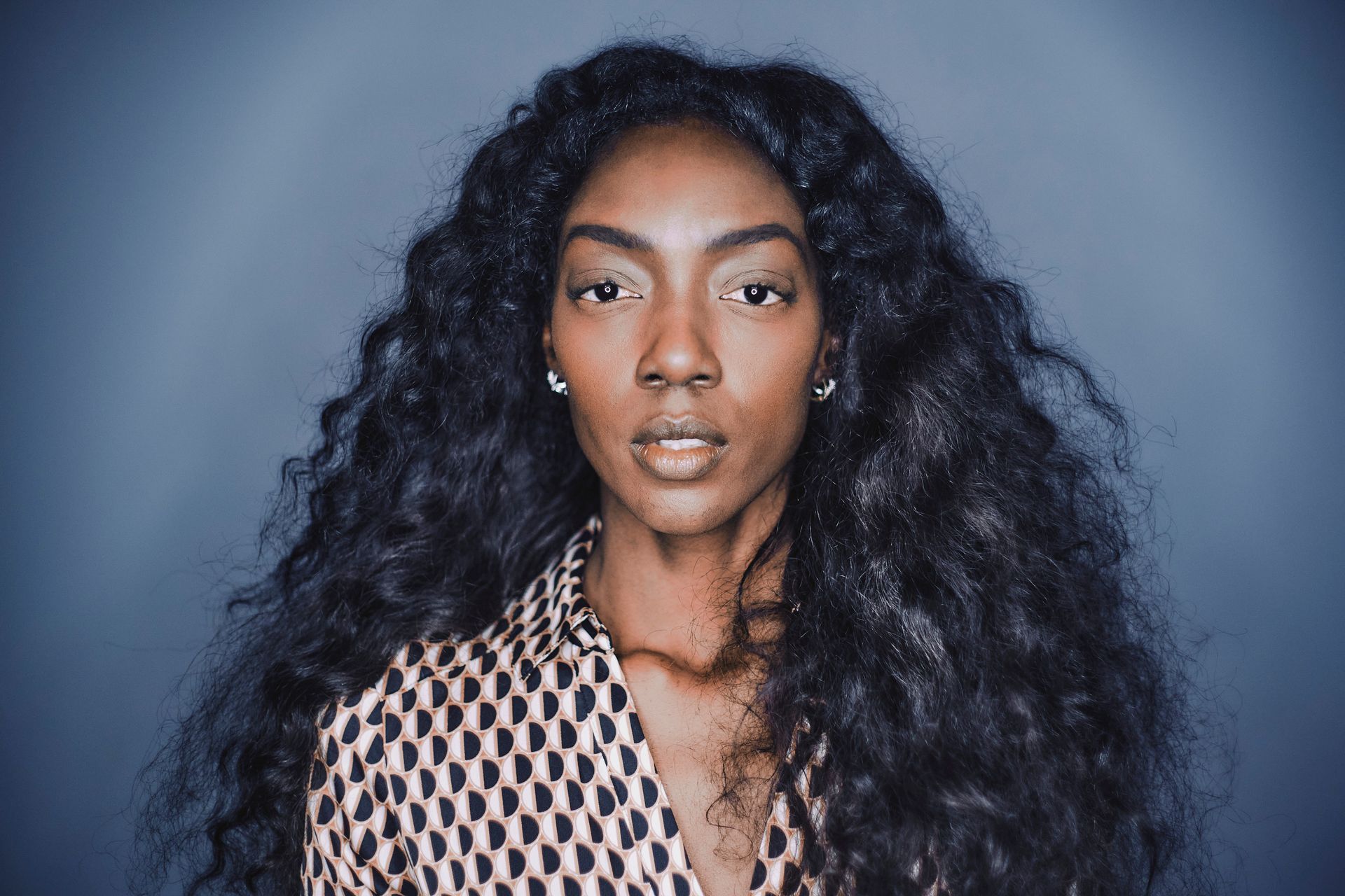 A woman with long curly hair is wearing a patterned shirt and earrings.