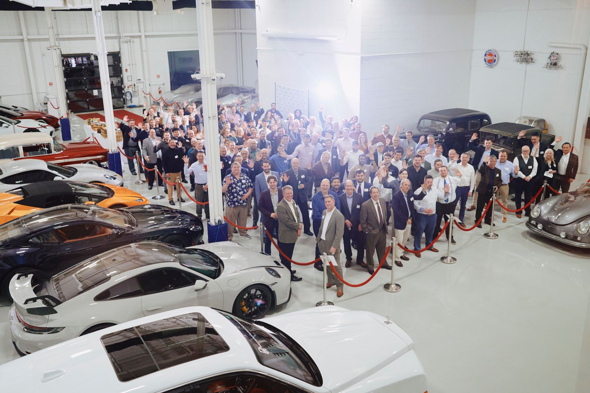 A large group of people are standing in a garage surrounded by cars.