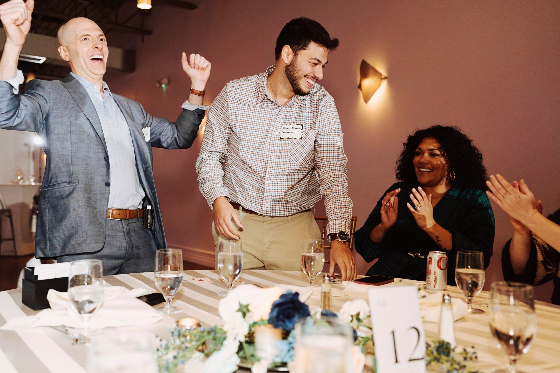 People celebrating at a dinner table, with one man standing and cheering while others clap and smile.