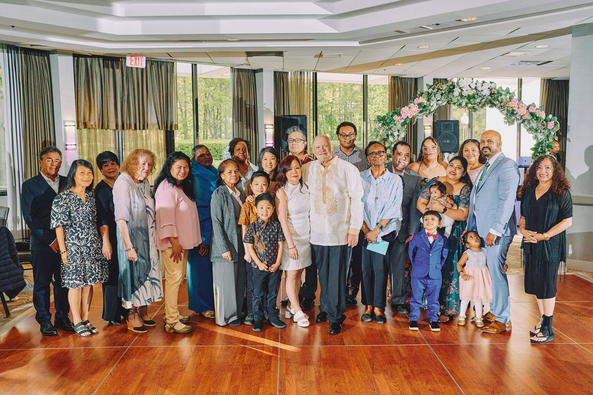 Group of people posing together in a bright indoor hall with wood flooring and large windows.