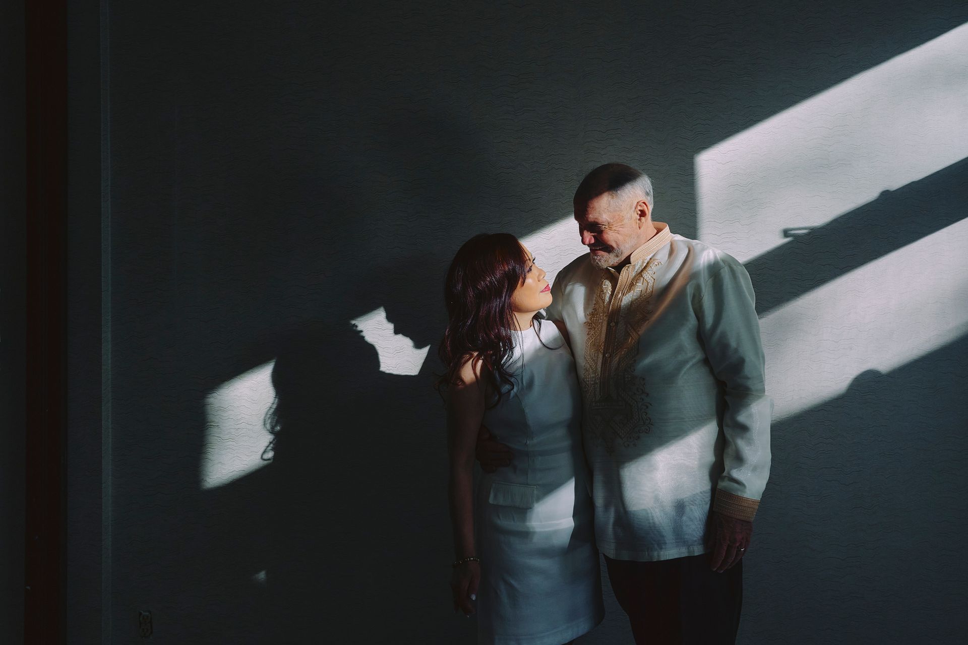 Two people standing close together in dim light, with bright window shadows on the wall behind them