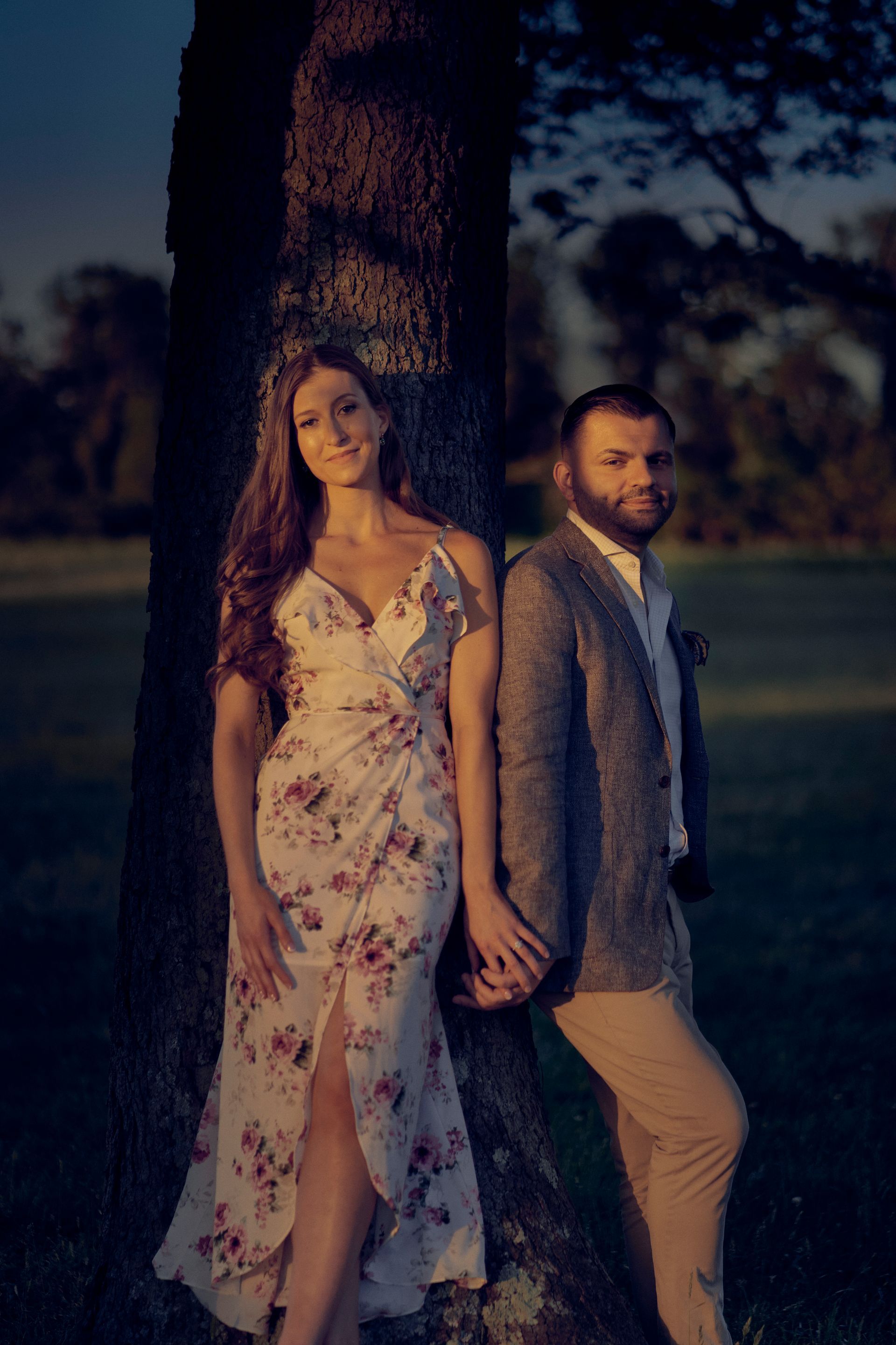A man and a woman are standing next to a tree holding hands.
