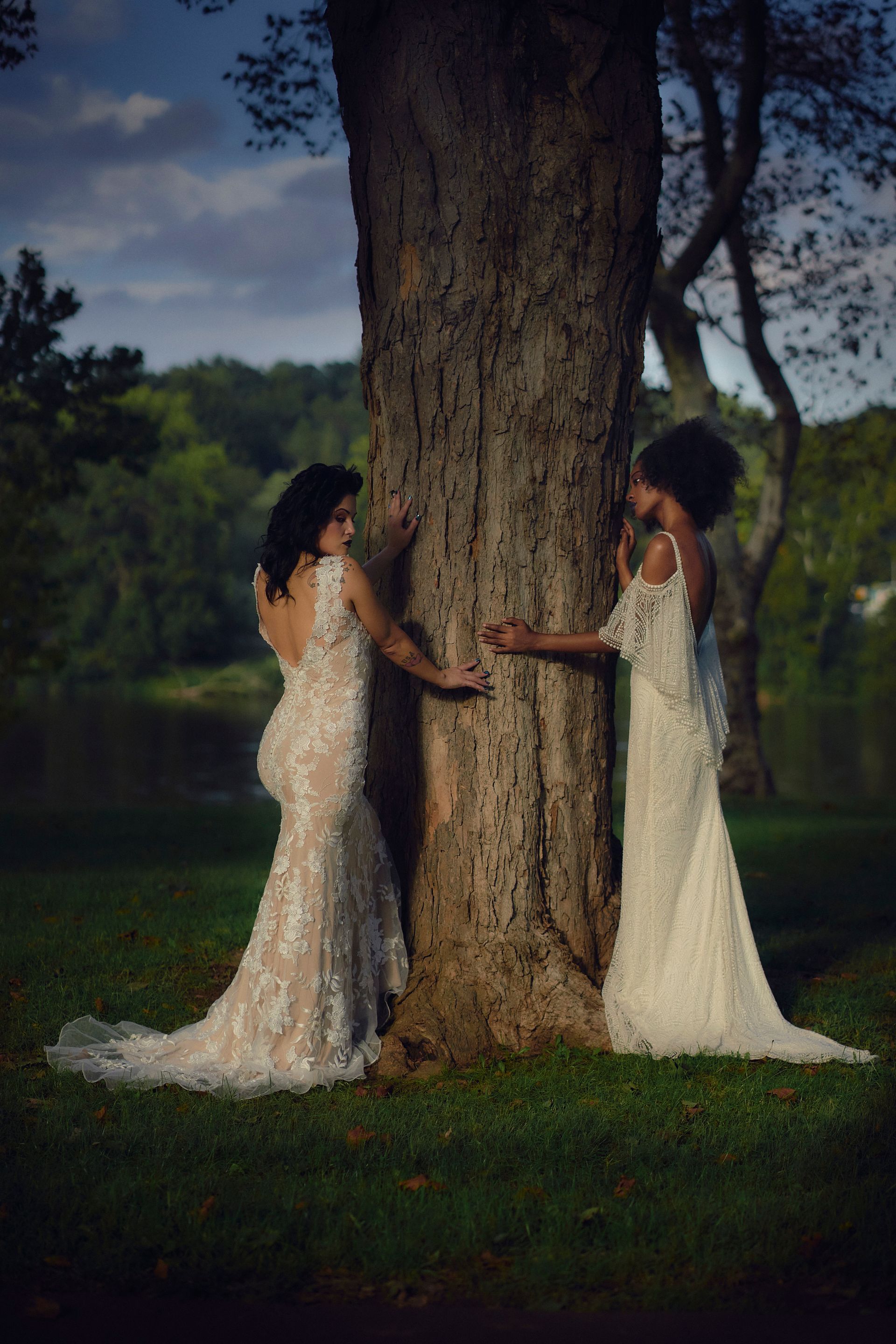 Two women in wedding dresses are hugging a tree.