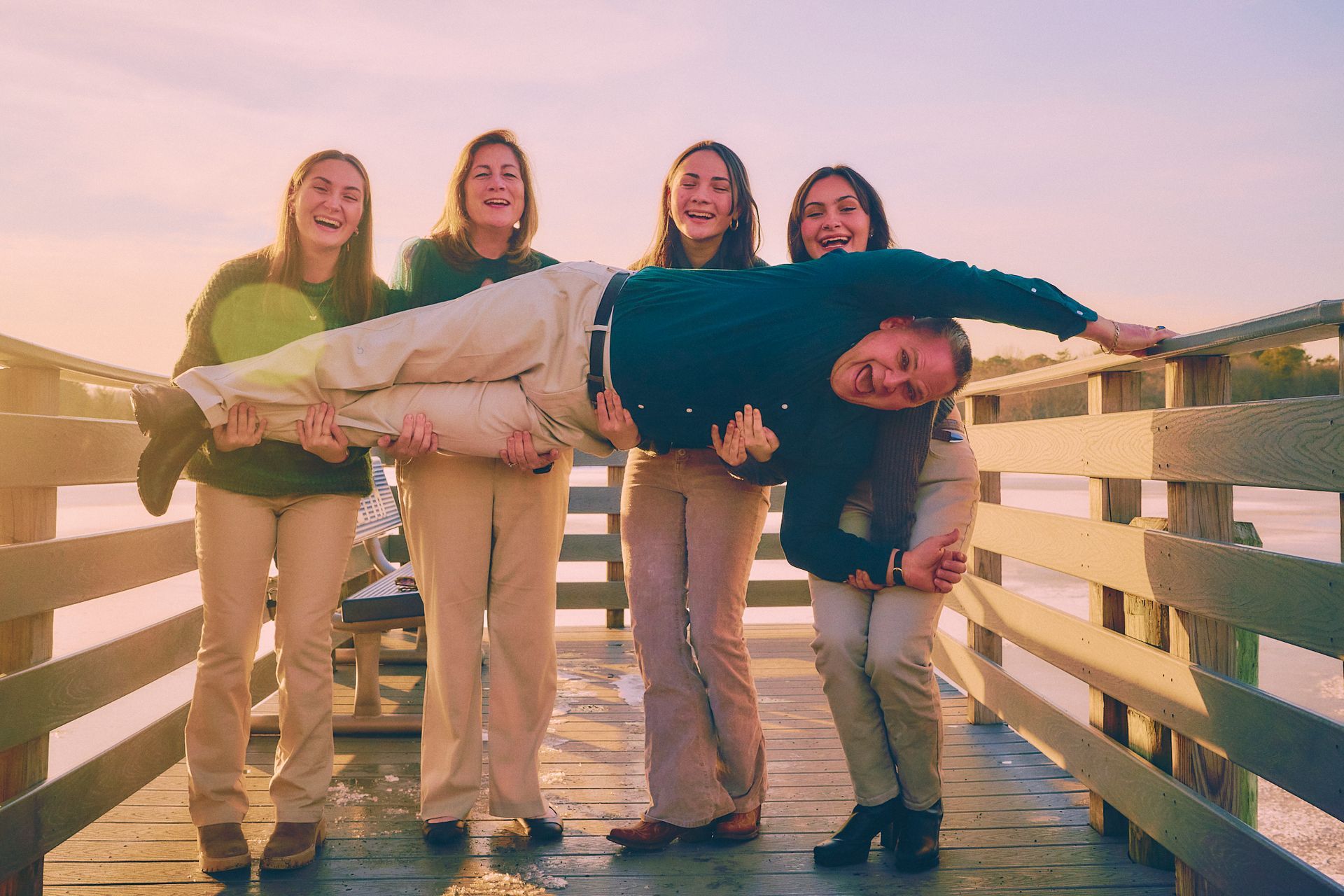 Four people lift one person on a wooden pier; all are smiling in the golden sunlight.